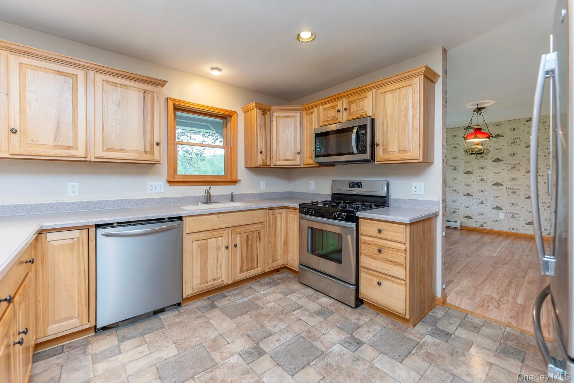 Kitchen with stainless steel appliances, a sink, light brown cabinetry, light countertops, and wallpapered walls Kitchen with stainless steel appliances, a sink, light brown cabinetry, light countertops, and wallpapered walls