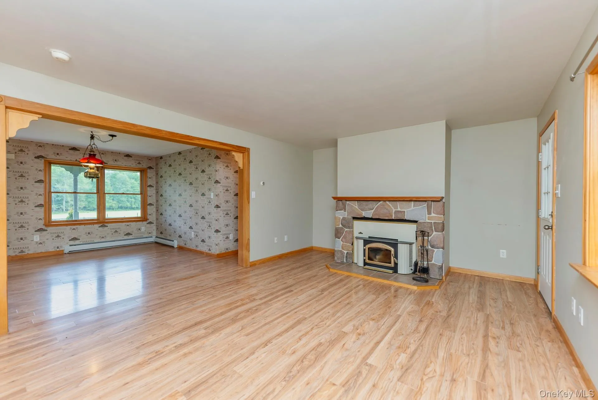 Unfurnished living room featuring baseboard heating, wood finished floors, baseboards, and wallpapered walls Unfurnished living room featuring baseboard heating, wood finished floors, baseboards, and wallpapered walls