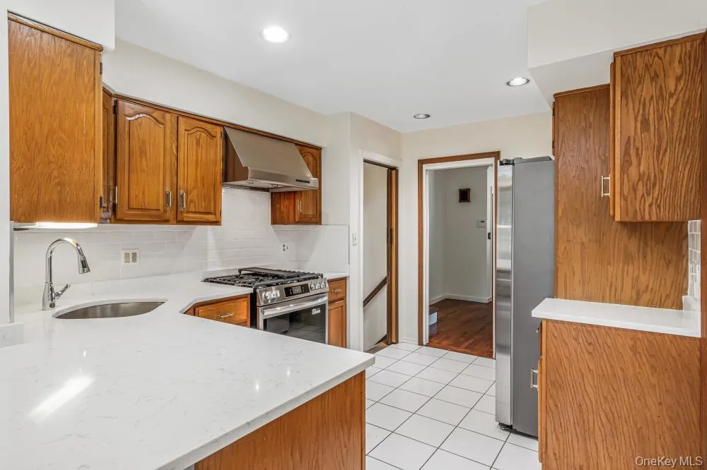 Kitchen featuring backsplash, brown cabinets, light tile patterned flooring, wall chimney exhaust hood, and recessed lighting Kitchen featuring backsplash, brown cabinets, light tile patterned flooring, wall chimney exhaust hood, and recessed lighting