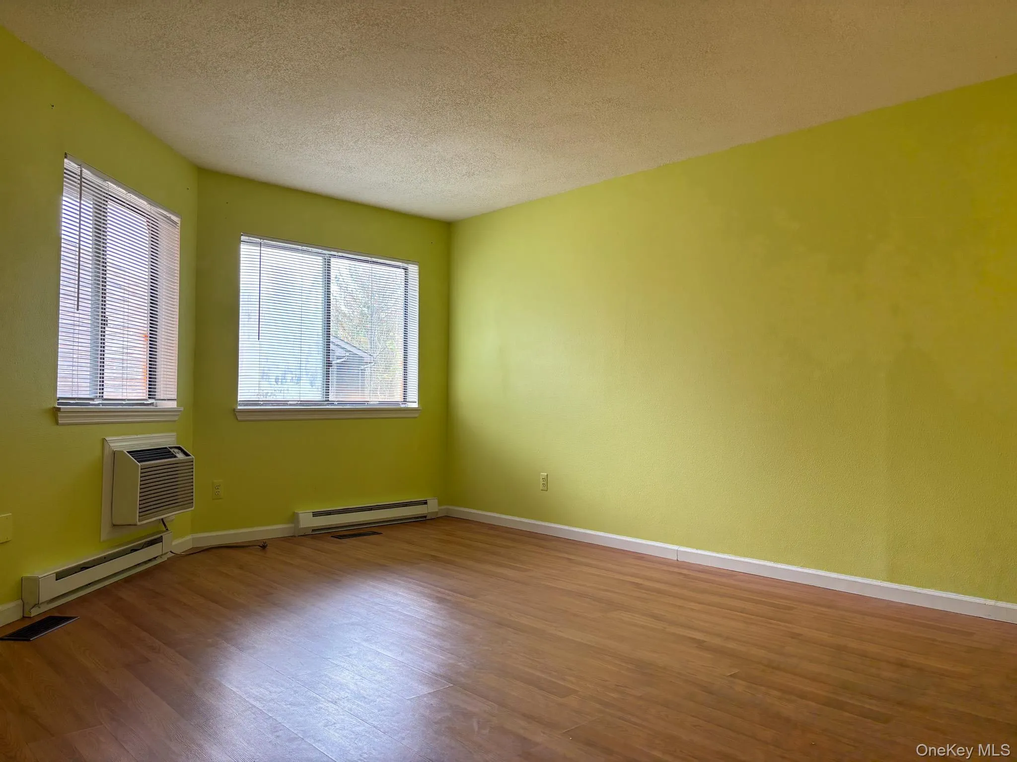 Primary beroom with a baseboard heating unit, wood finished floors, a textured ceiling, baseboards, and a wall mounted air conditioner Primary beroom with a baseboard heating unit, wood finished floors, a textured ceiling, baseboards, and a wall mounted air conditioner