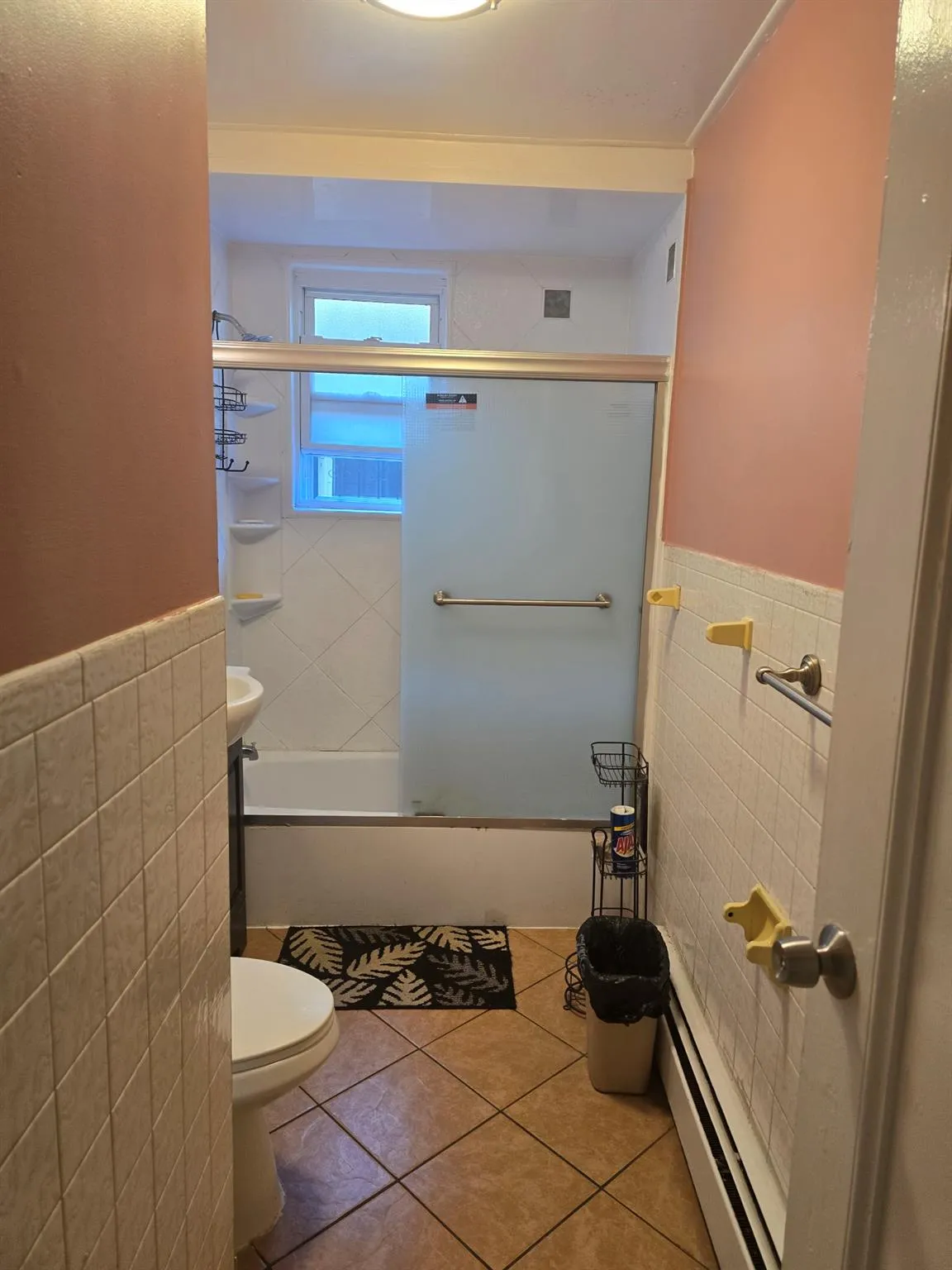 Full bath featuring tile walls, baseboard heating, toilet, bath / shower combo with glass door, and wainscoting Full bath featuring tile walls, baseboard heating, toilet, bath / shower combo with glass door, and wainscoting