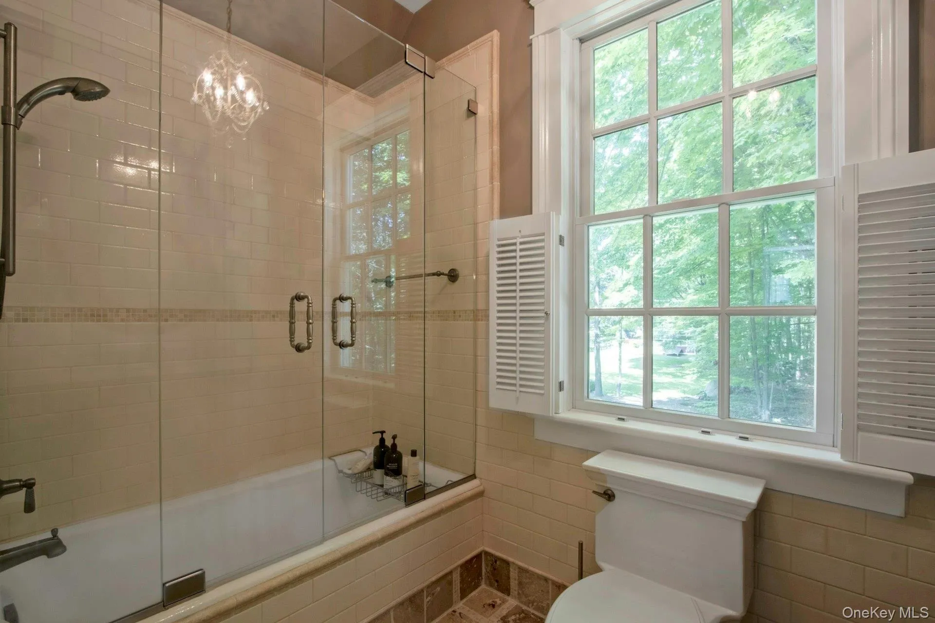 Bathroom with healthy amount of natural light, shower / bath combination with glass door, toilet, tile walls, and a chandelier Bathroom with healthy amount of natural light, shower / bath combination with glass door, toilet, tile walls, and a chandelier