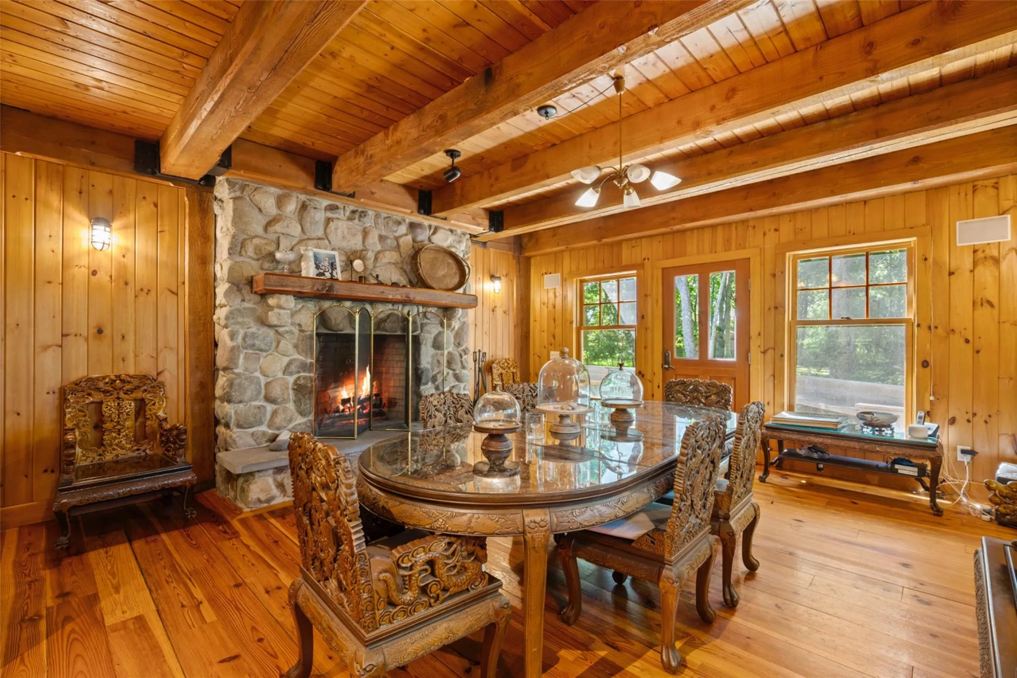 Dining space featuring light wood-style flooring, wooden ceiling with exposed beams, wood walls, a stone fireplace, and a ceiling fan Dining space featuring light wood-style flooring, wooden ceiling with exposed beams, wood walls, a stone fireplace, and a ceiling fan