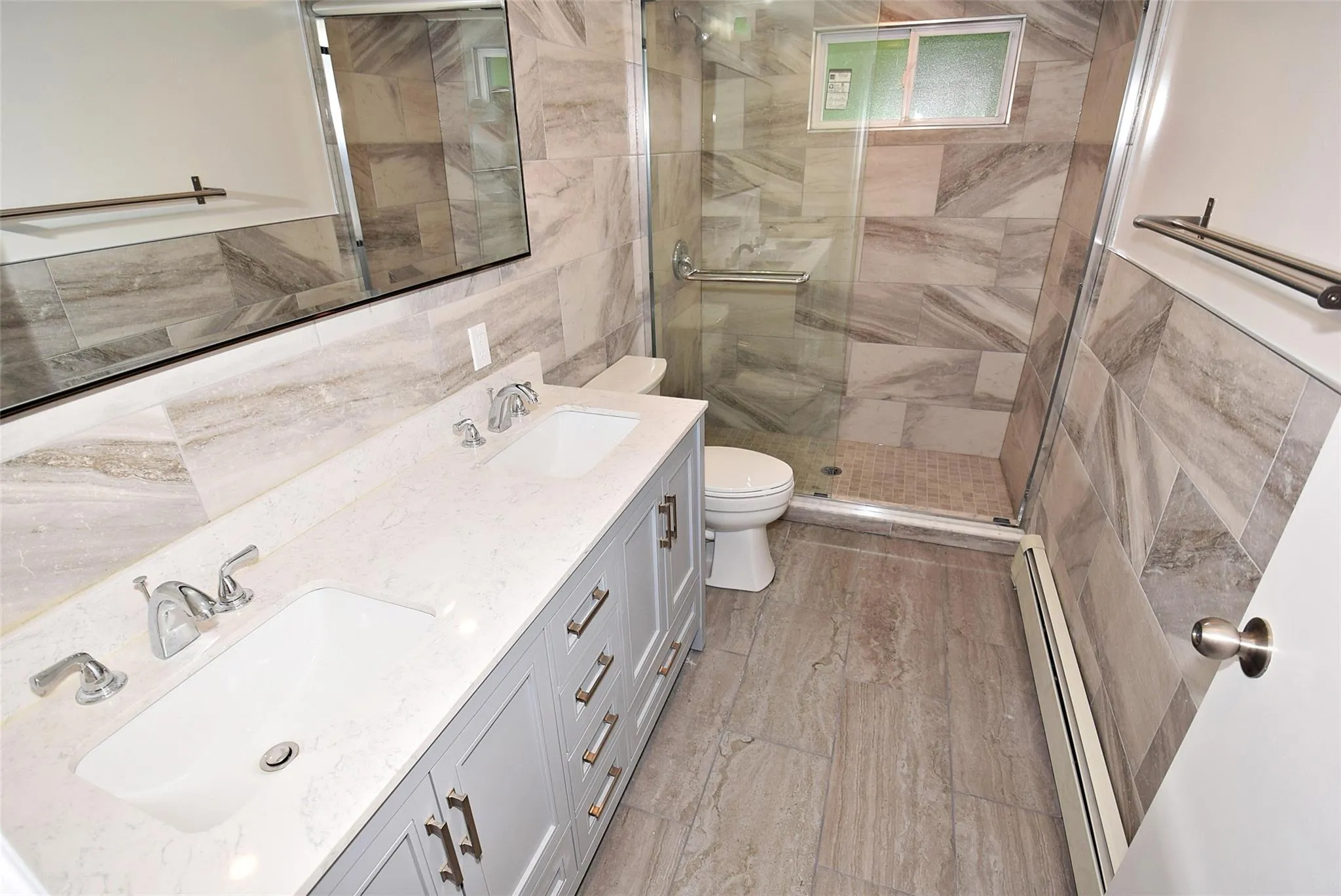 Bathroom with baseboard heating, toilet, double vanity, a shower stall, and tile walls Bathroom with baseboard heating, toilet, double vanity, a shower stall, and tile walls