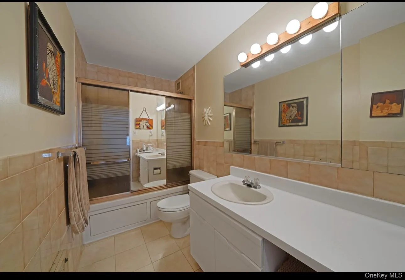 Bathroom with tile walls, bath / shower combo with glass door, vanity, light tile patterned flooring, and a wainscoted wall Bathroom with tile walls, bath / shower combo with glass door, vanity, light tile patterned flooring, and a wainscoted wall
