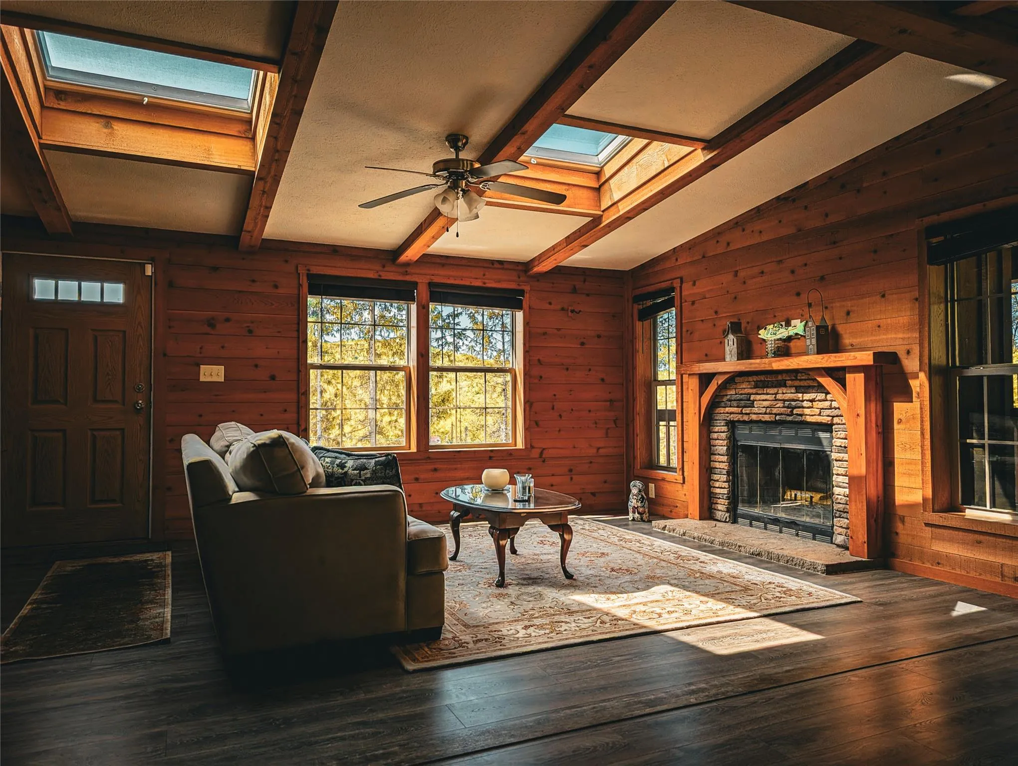 Living area with a skylight, a brick fireplace, wood walls, a ceiling fan, and wood-type flooring Living area with a skylight, a brick fireplace, wood walls, a ceiling fan, and wood-type flooring