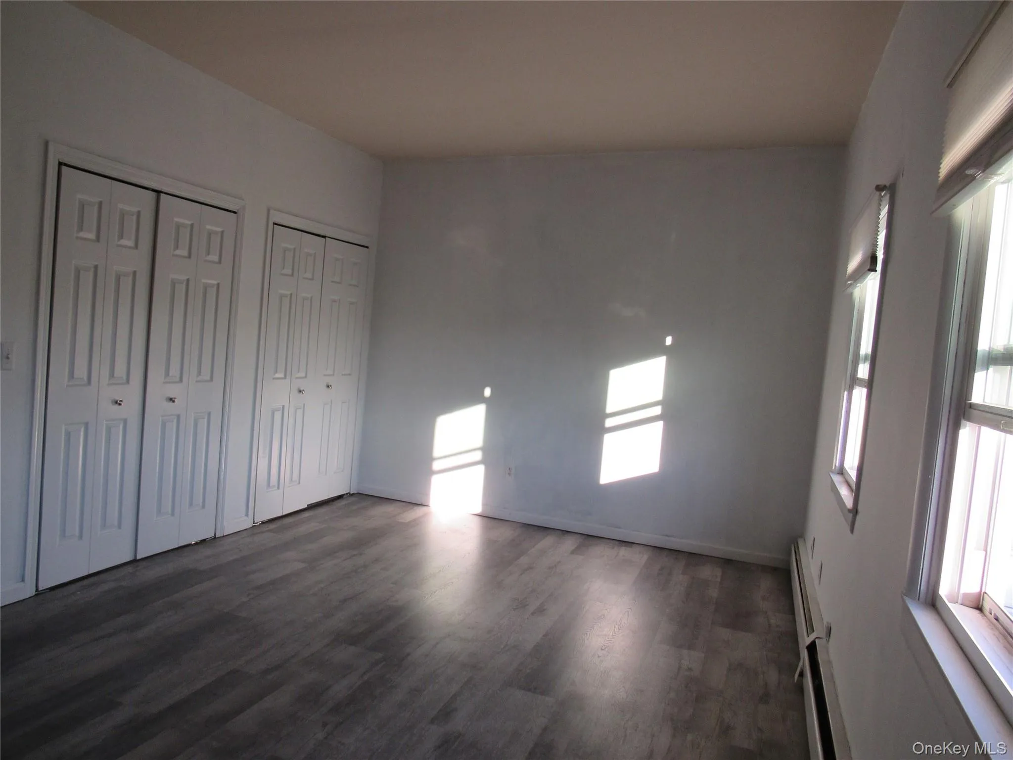 Unfurnished bedroom featuring two closets, wood finished floors, multiple windows, and a baseboard radiator Unfurnished bedroom featuring two closets, wood finished floors, multiple windows, and a baseboard radiator