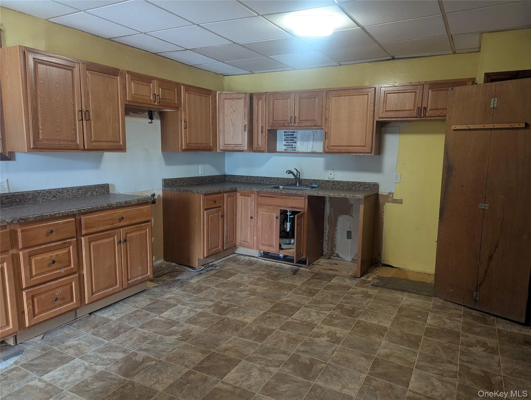 Kitchen with a drop ceiling, dark countertops, brown cabinets, and stone finish floors Kitchen with a drop ceiling, dark countertops, brown cabinets, and stone finish floors