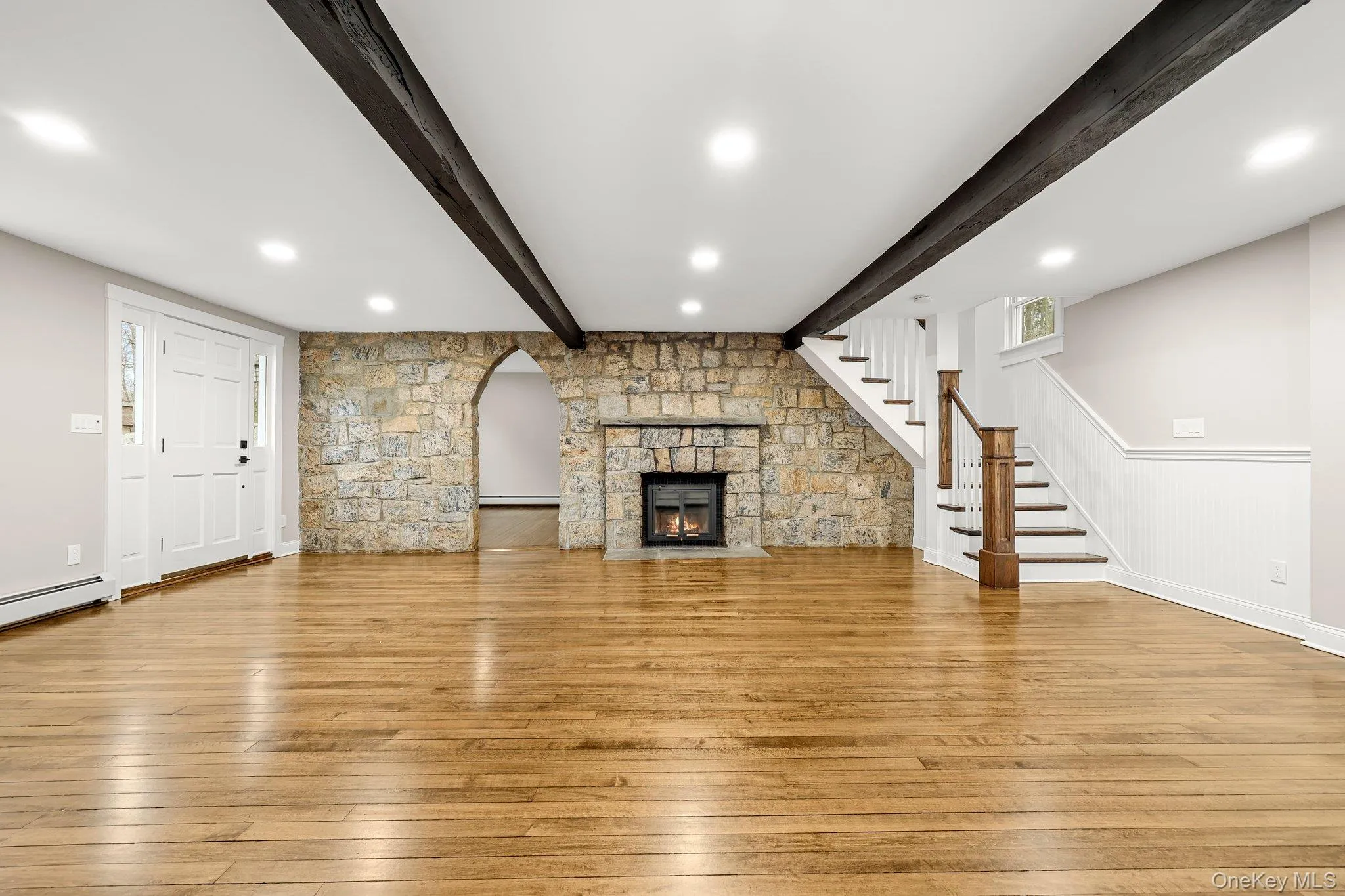Living room with beam ceiling, light wood finished floors, stairs, a stone fireplace, and a baseboard radiator Living room with beam ceiling, light wood finished floors, stairs, a stone fireplace, and a baseboard radiator