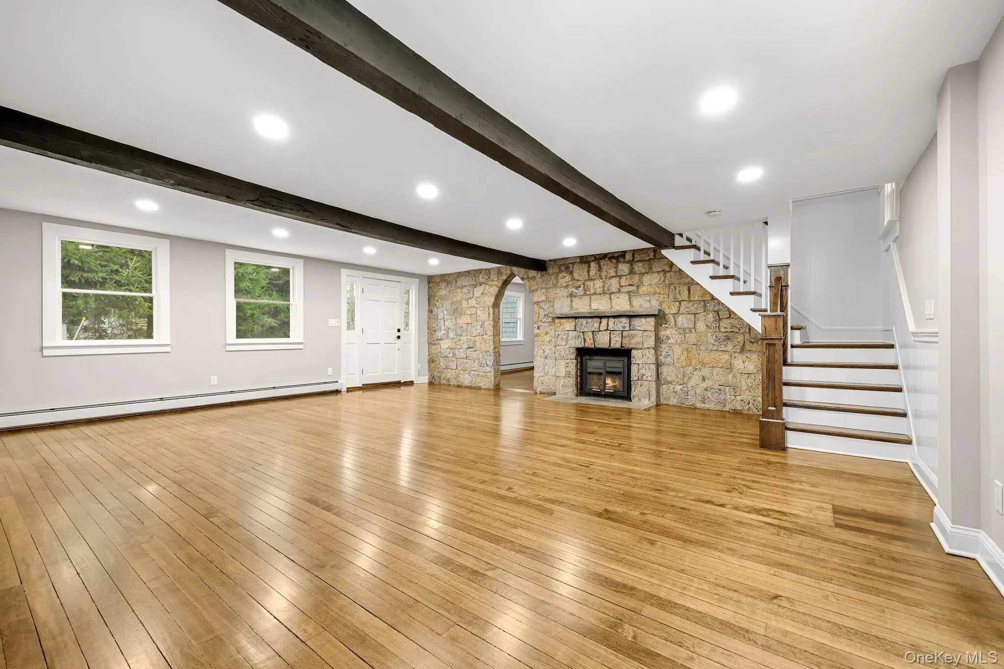 Living room featuring baseboard heating, beamed ceiling, stairs, wood finished floors, and a stone fireplace Living room featuring baseboard heating, beamed ceiling, stairs, wood finished floors, and a stone fireplace
