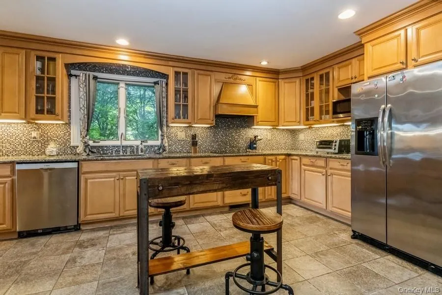 Kitchen featuring appliances with stainless steel finishes, custom exhaust hood, decorative backsplash, dark stone countertops, and glass insert cabinets Kitchen featuring appliances with stainless steel finishes, custom exhaust hood, decorative backsplash, dark stone countertops, and glass insert cabinets