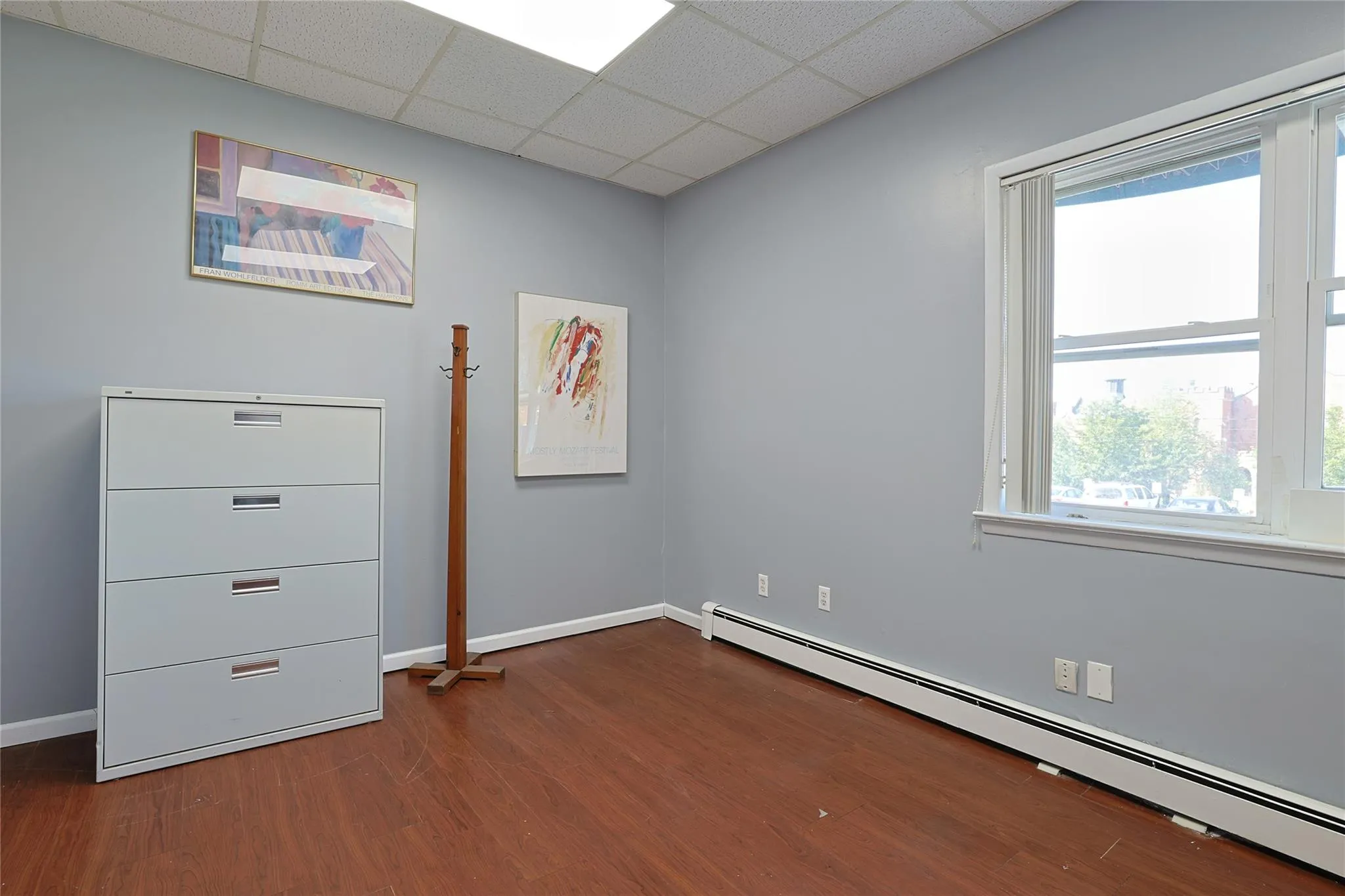 Empty room with a baseboard heating unit, dark wood-type flooring, healthy amount of natural light, and a drop ceiling Empty room with a baseboard heating unit, dark wood-type flooring, healthy amount of natural light, and a drop ceiling