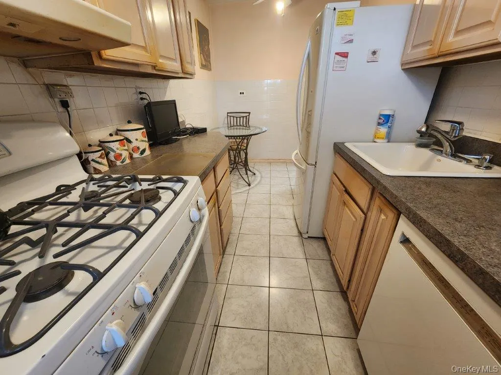 Kitchen with white appliances, under cabinet range hood, tasteful backsplash, light tile patterned floors, and dark countertops Kitchen with white appliances, under cabinet range hood, tasteful backsplash, light tile patterned floors, and dark countertops