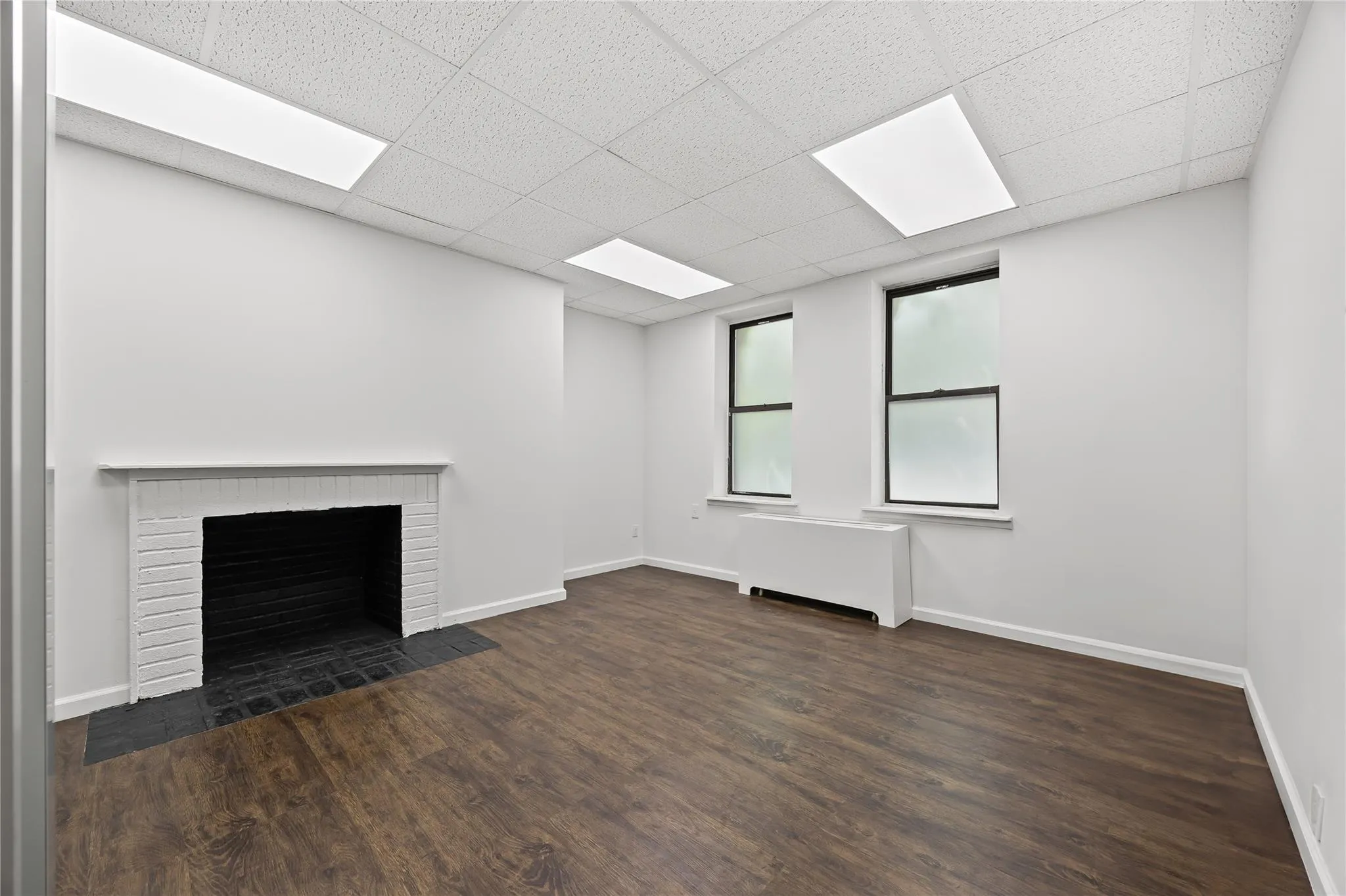 Unfurnished living room with wood finished floors, radiator heating unit, a brick fireplace, and a paneled ceiling Unfurnished living room with wood finished floors, radiator heating unit, a brick fireplace, and a paneled ceiling