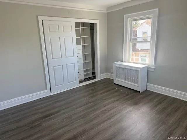 Unfurnished bedroom with radiator, dark wood-style floors, crown molding, and a closet Unfurnished bedroom with radiator, dark wood-style floors, crown molding, and a closet