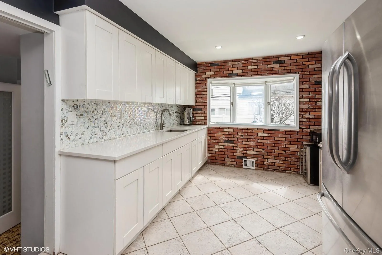 Kitchen with freestanding refrigerator, white cabinetry, light tile patterned floors, brick wall, and light stone countertops Kitchen with freestanding refrigerator, white cabinetry, light tile patterned floors, brick wall, and light stone countertops