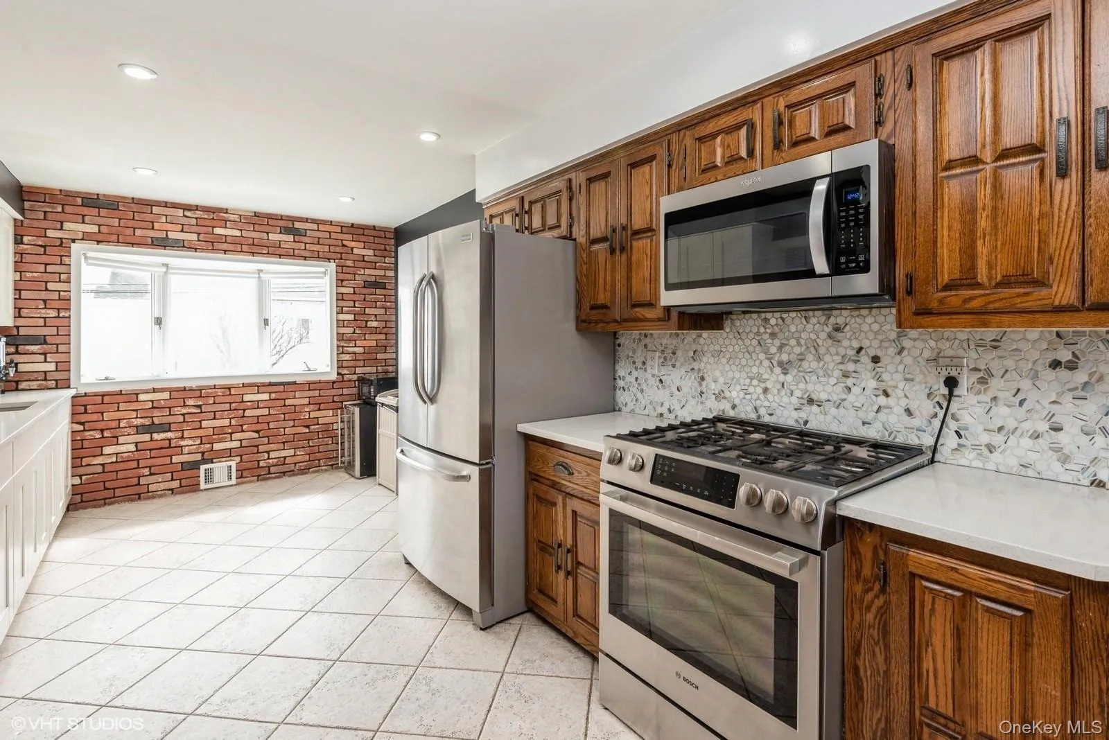 Kitchen featuring stainless steel appliances, brick wall, light tile patterned flooring, brown cabinetry, and tasteful backsplash Kitchen featuring stainless steel appliances, brick wall, light tile patterned flooring, brown cabinetry, and tasteful backsplash