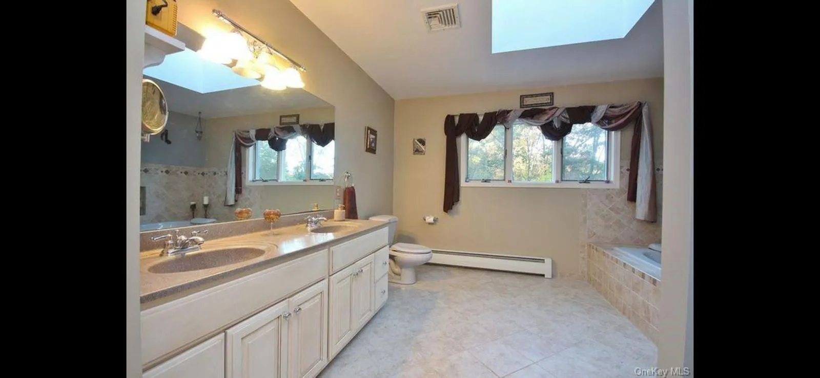 Full bathroom featuring a baseboard heating unit, double vanity, tiled tub, a skylight, and tile patterned floors Full bathroom featuring a baseboard heating unit, double vanity, tiled tub, a skylight, and tile patterned floors