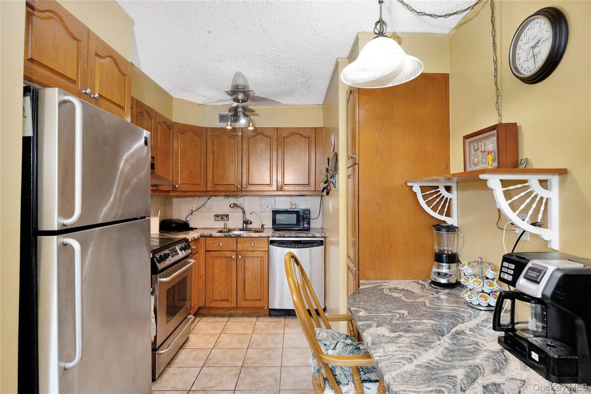 Kitchen featuring appliances with stainless steel finishes, ceiling fan, brown cabinets, light tile patterned floors, and a textured ceiling Kitchen featuring appliances with stainless steel finishes, ceiling fan, brown cabinets, light tile patterned floors, and a textured ceiling