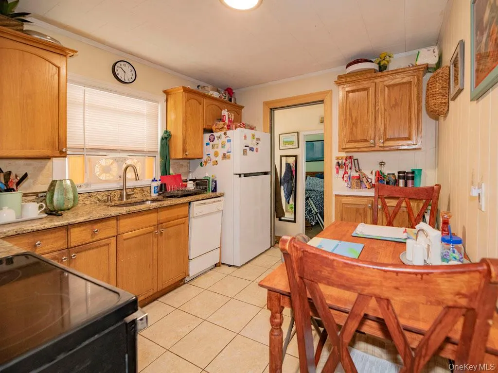 Kitchen featuring white appliances, ornamental molding, light tile patterned flooring, light stone counters, and brown cabinetry Kitchen featuring white appliances, ornamental molding, light tile patterned flooring, light stone counters, and brown cabinetry