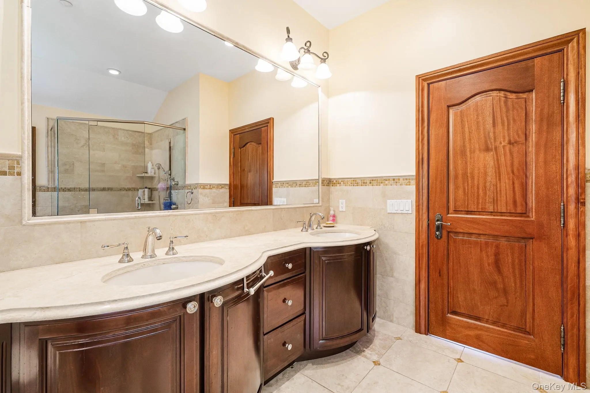 Full bathroom with double vanity, tile walls, tile patterned floors, tiled shower, and wainscoting Full bathroom with double vanity, tile walls, tile patterned floors, tiled shower, and wainscoting
