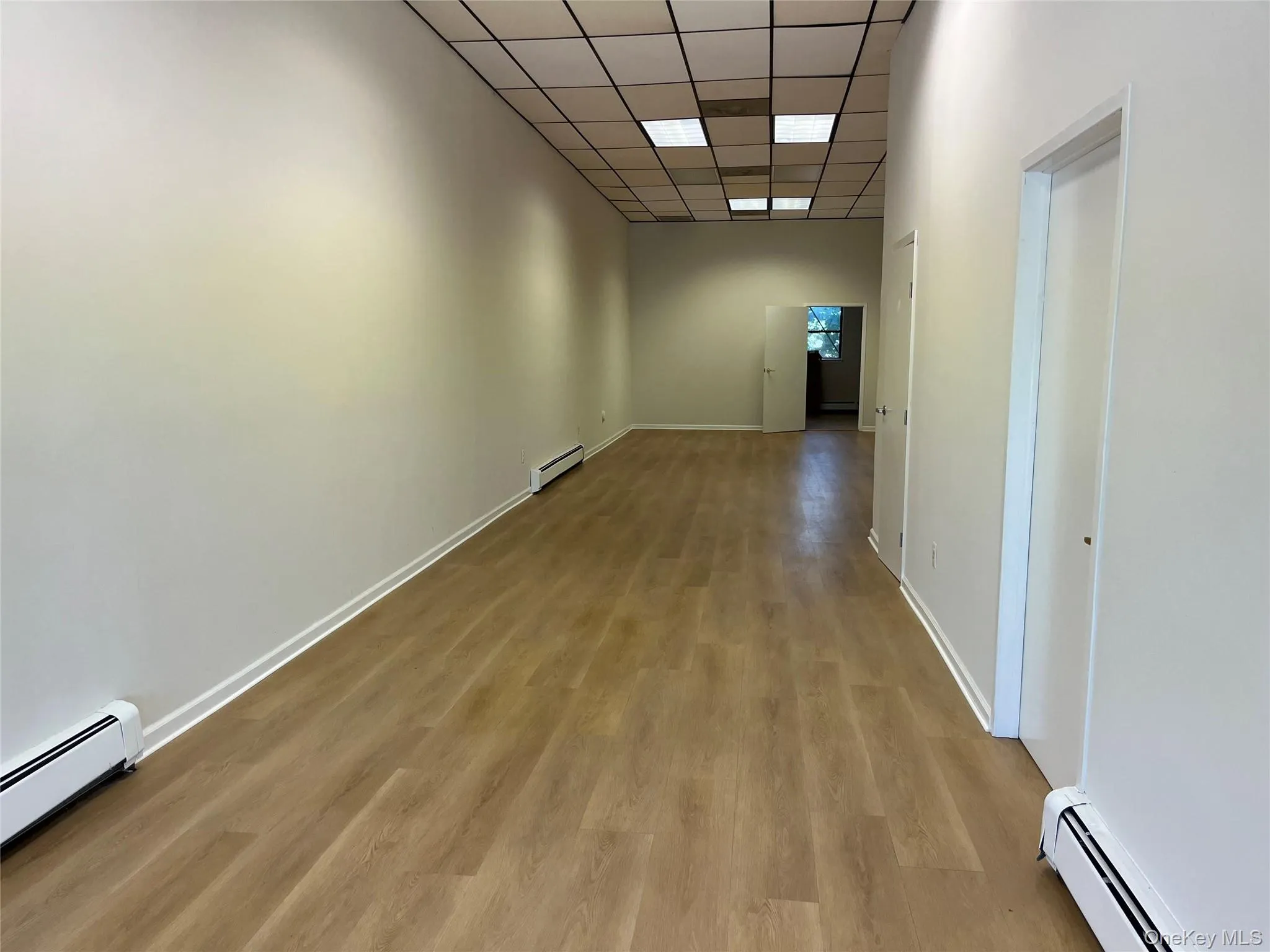 Hall featuring baseboard heating, a drop ceiling, and light wood finished floors Hall featuring baseboard heating, a drop ceiling, and light wood finished floors