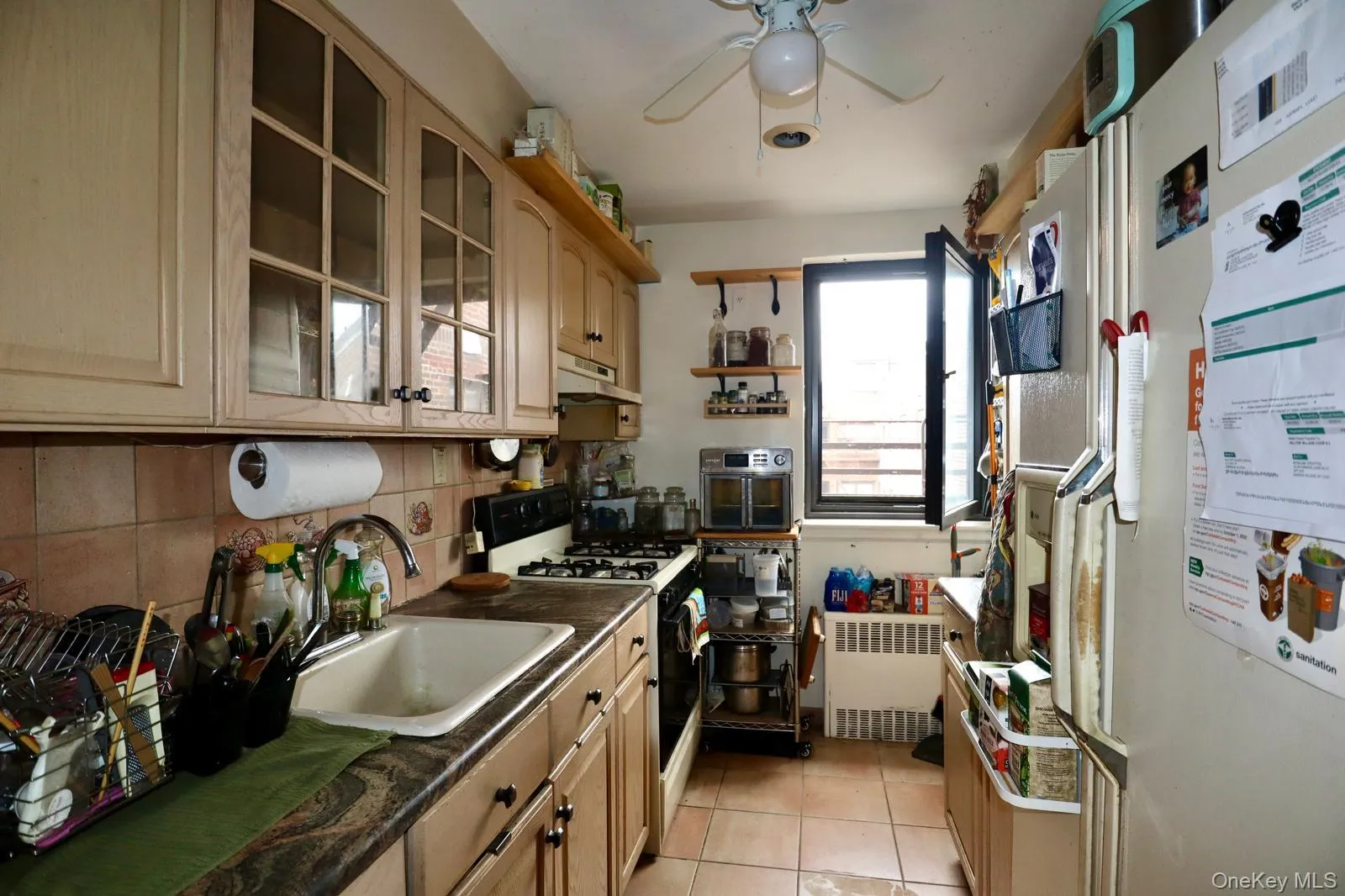 Kitchen featuring white appliances, radiator, ceiling fan, glass insert cabinets, and dark countertops Kitchen featuring white appliances, radiator, ceiling fan, glass insert cabinets, and dark countertops