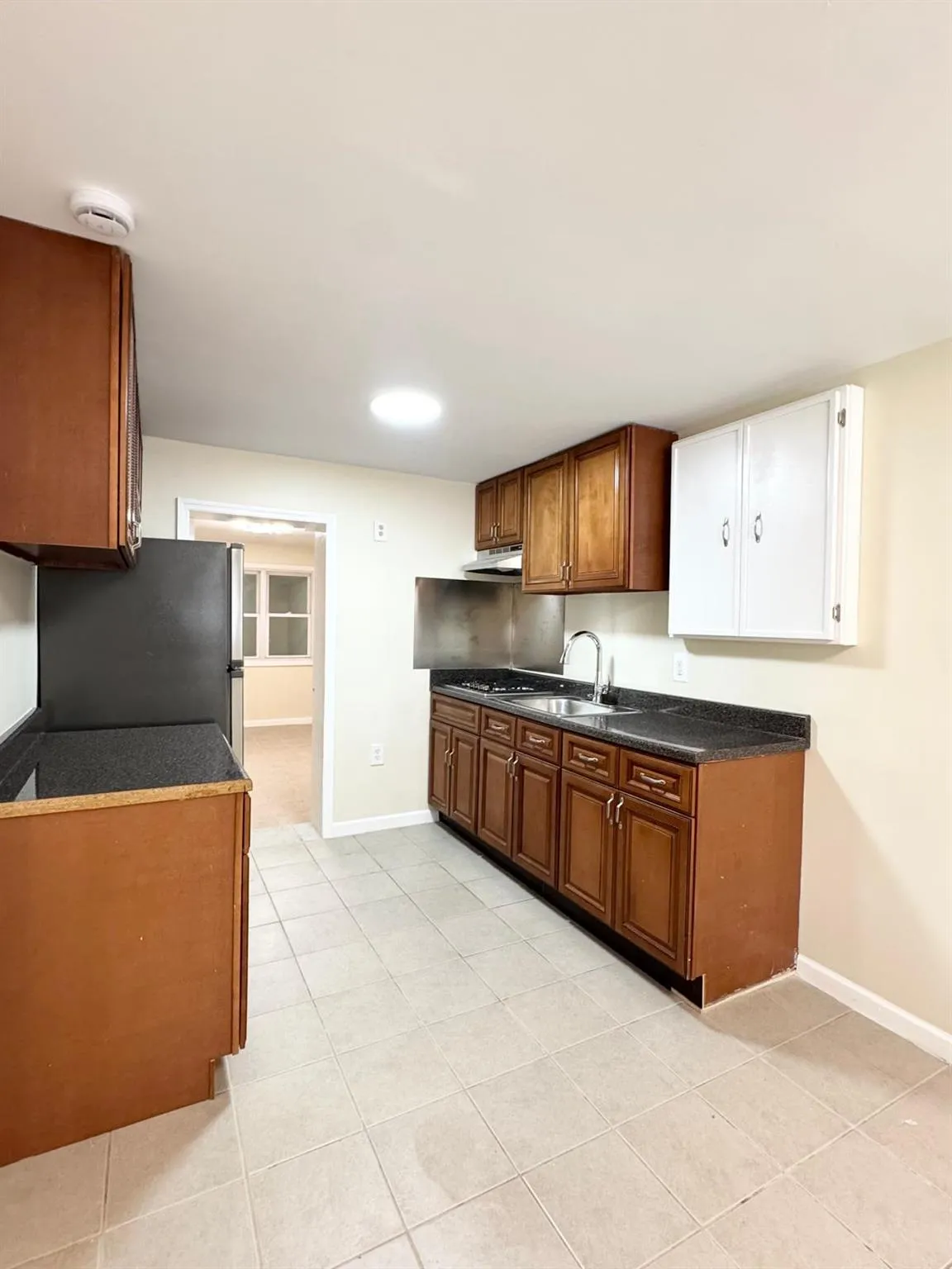 Kitchen with freestanding refrigerator, under cabinet range hood, light tile patterned floors, dark countertops, and brown cabinetry Kitchen with freestanding refrigerator, under cabinet range hood, light tile patterned floors, dark countertops, and brown cabinetry