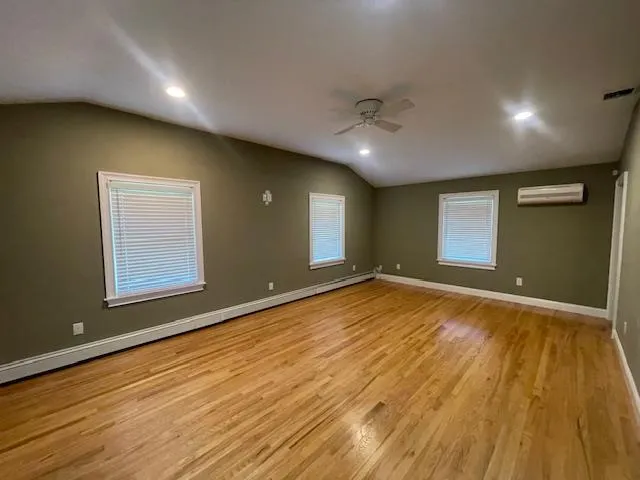 Empty room featuring lofted ceiling, an AC wall unit, ceiling fan, wood finished floors, and a baseboard heating unit Empty room featuring lofted ceiling, an AC wall unit, ceiling fan, wood finished floors, and a baseboard heating unit