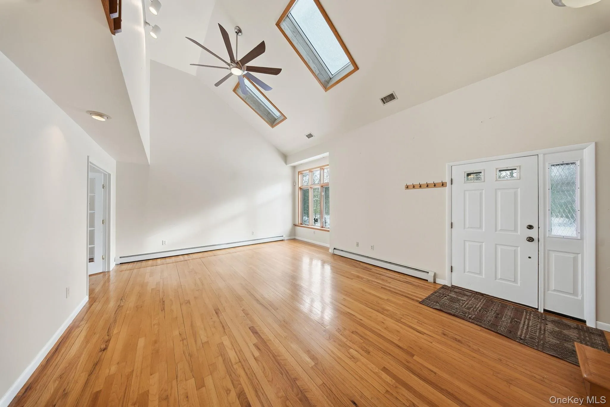 Unfurnished living room featuring high vaulted ceiling, a skylight, baseboard heating, light wood-style floors, and ceiling fan Unfurnished living room featuring high vaulted ceiling, a skylight, baseboard heating, light wood-style floors, and ceiling fan