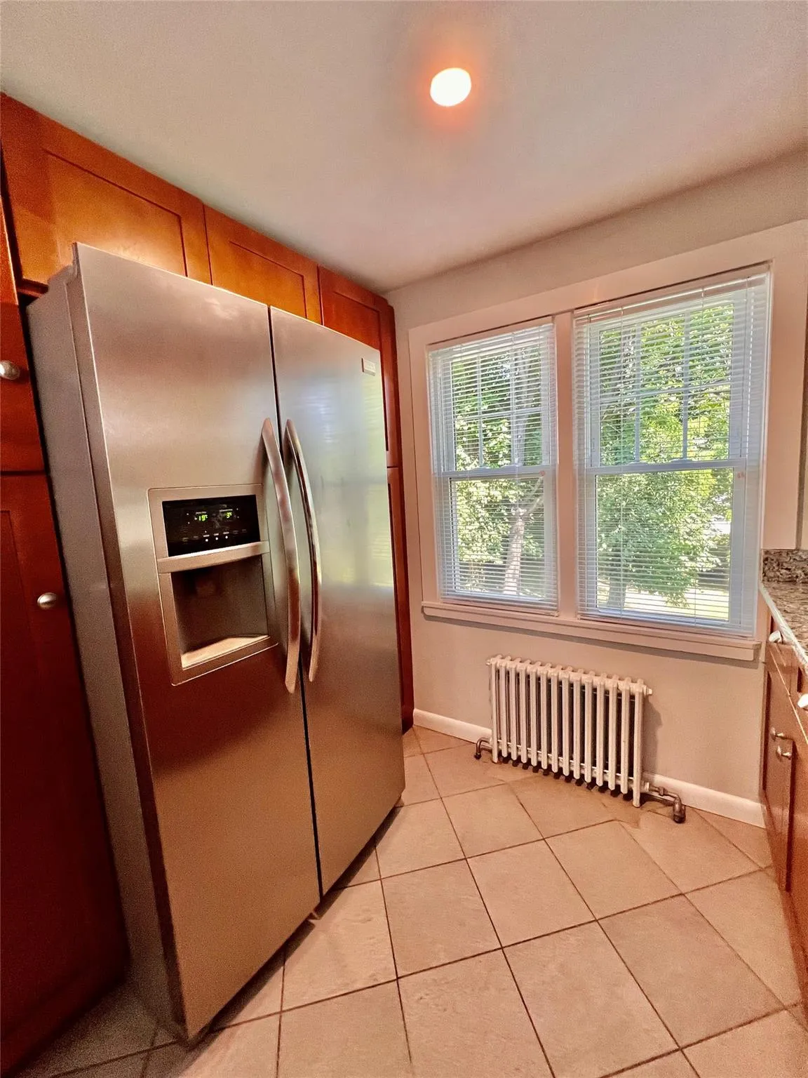 Kitchen featuring stainless steel refrigerator with ice dispenser, radiator, light tile patterned floors, and brown cabinets Kitchen featuring stainless steel refrigerator with ice dispenser, radiator, light tile patterned floors, and brown cabinets