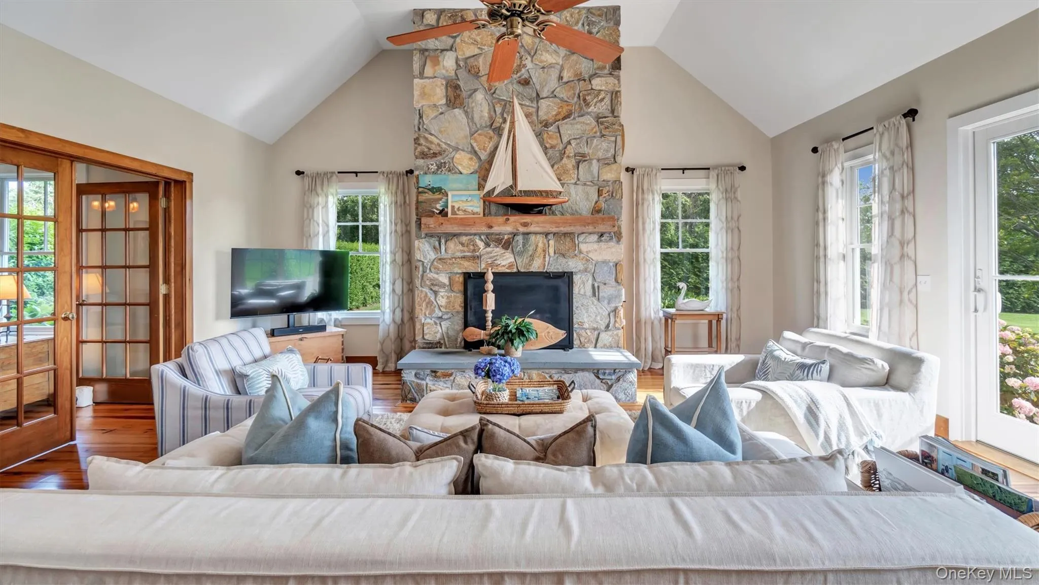Living room with healthy amount of natural light, a ceiling fan, wood finished floors, a stone fireplace, and high vaulted ceiling Living room with healthy amount of natural light, a ceiling fan, wood finished floors, a stone fireplace, and high vaulted ceiling