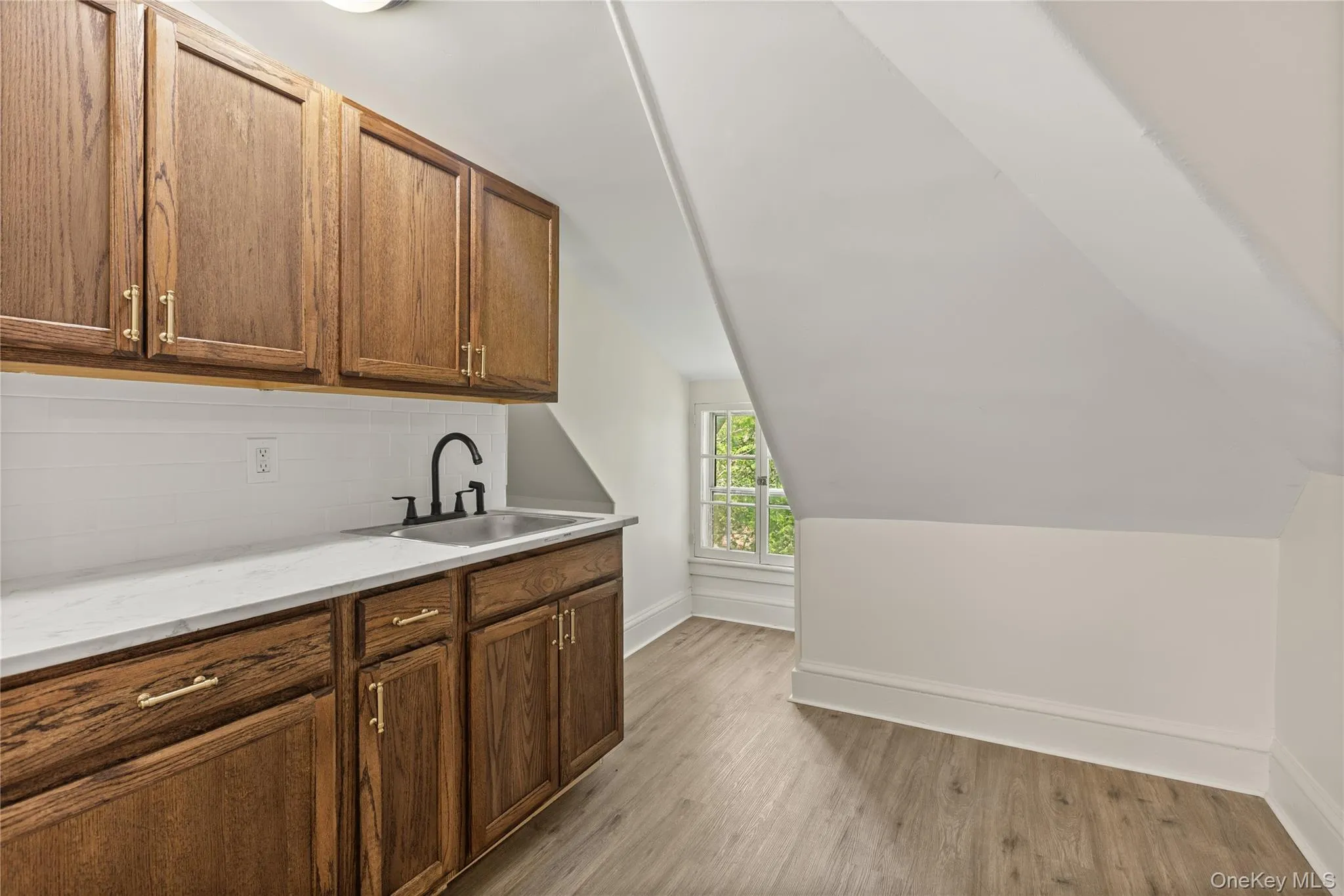 Bar featuring light wood finished floors, decorative backsplash, vaulted ceiling, brown cabinetry, and light stone countertops Bar featuring light wood finished floors, decorative backsplash, vaulted ceiling, brown cabinetry, and light stone countertops