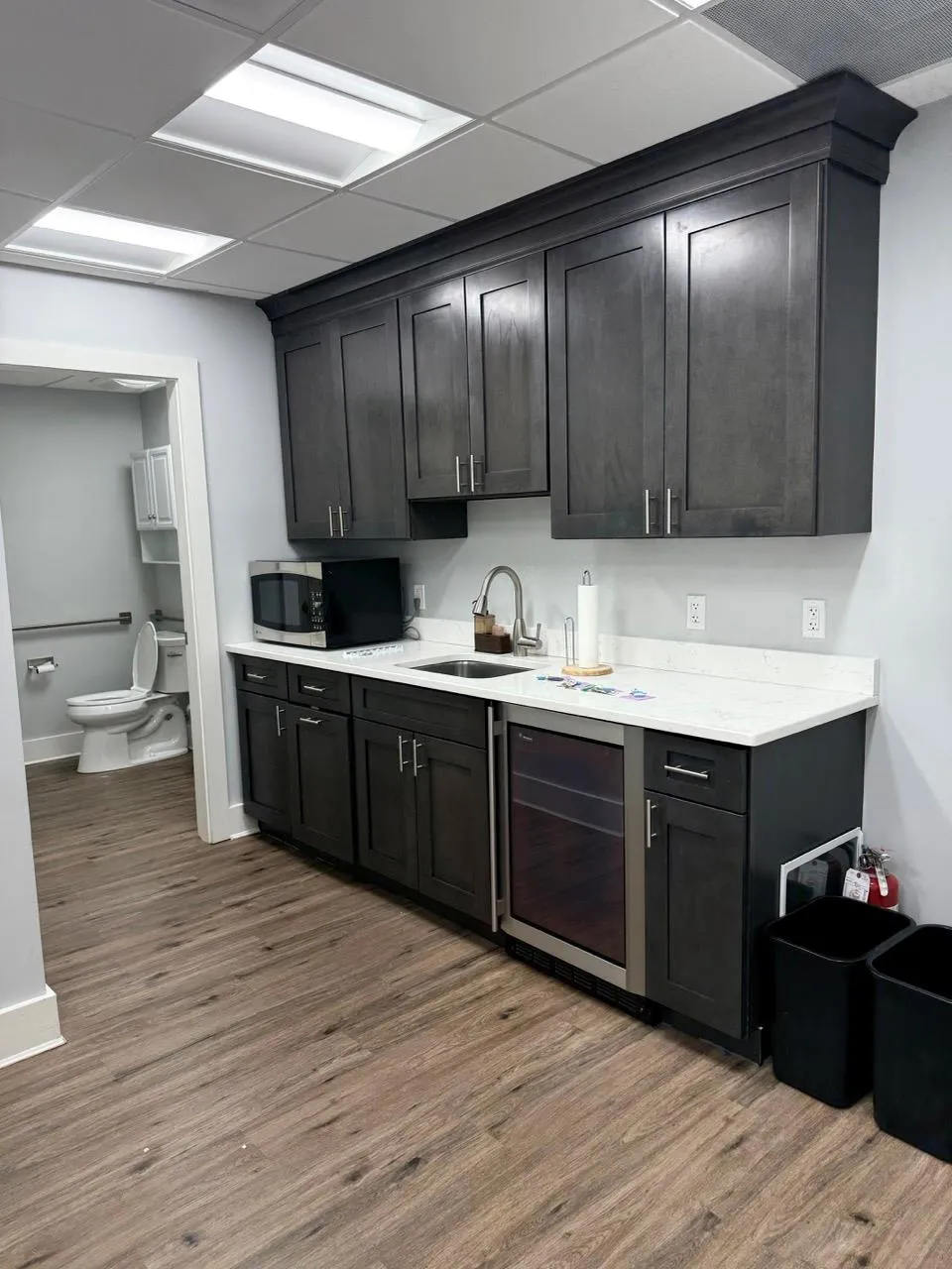 Kitchen with light countertops, stainless steel microwave, wood finished floors, wine cooler, and dark cabinetry Kitchen with light countertops, stainless steel microwave, wood finished floors, wine cooler, and dark cabinetry