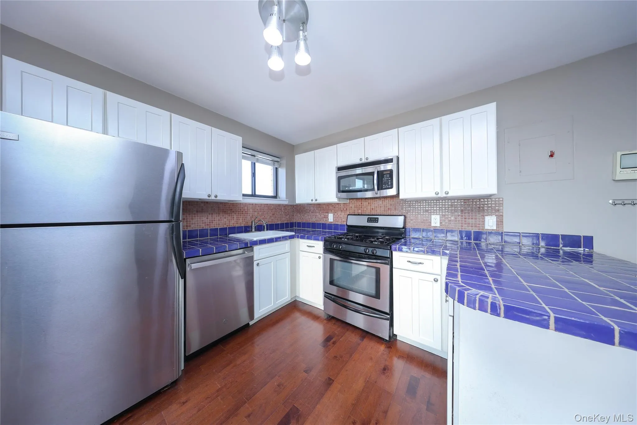 Kitchen with tile counters, stainless steel appliances, white cabinets, dark wood-type flooring, and decorative backsplash Kitchen with tile counters, stainless steel appliances, white cabinets, dark wood-type flooring, and decorative backsplash