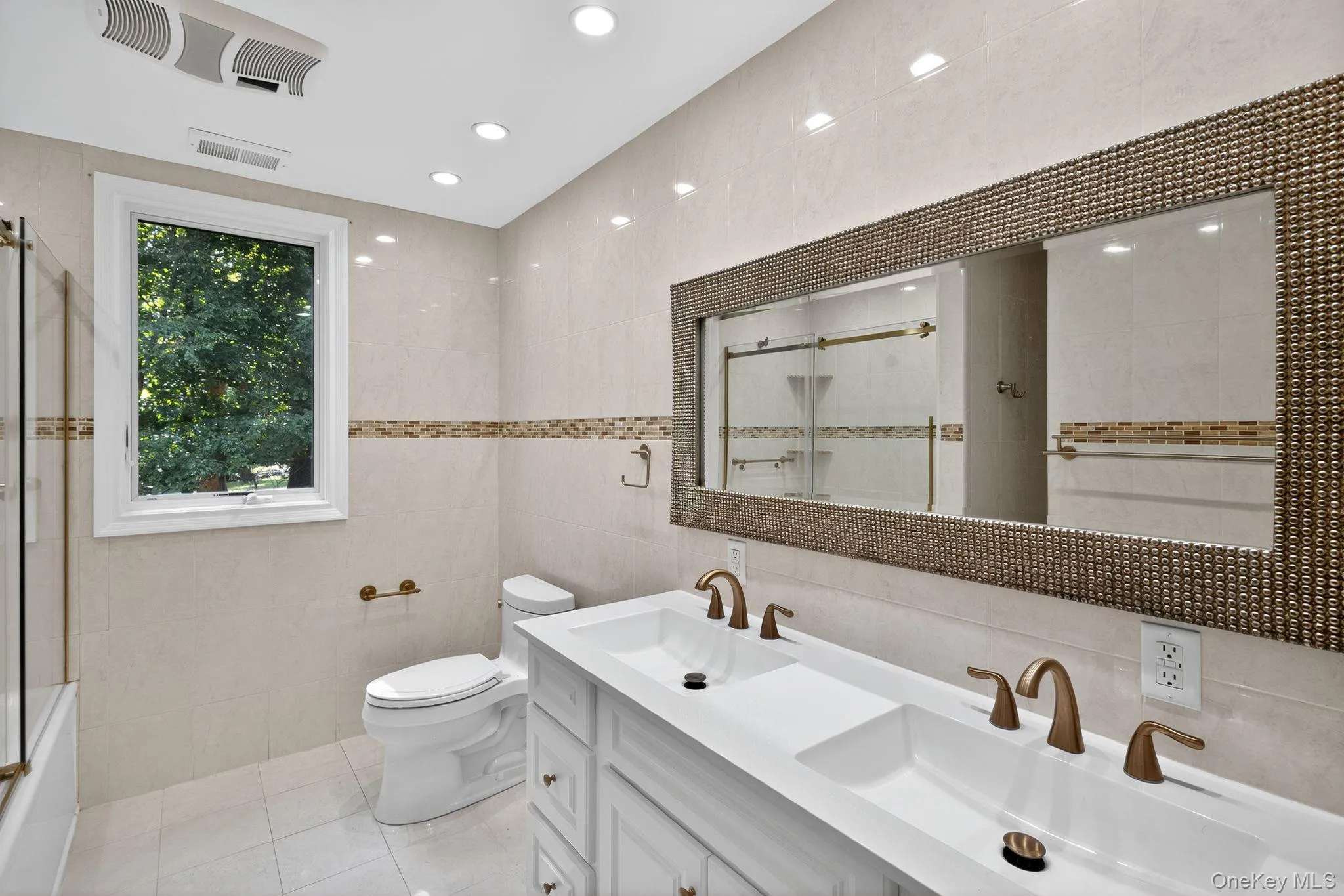 Bathroom featuring tile walls, double vanity, light tile patterned floors, recessed lighting, and bath / shower combo with glass door Bathroom featuring tile walls, double vanity, light tile patterned floors, recessed lighting, and bath / shower combo with glass door