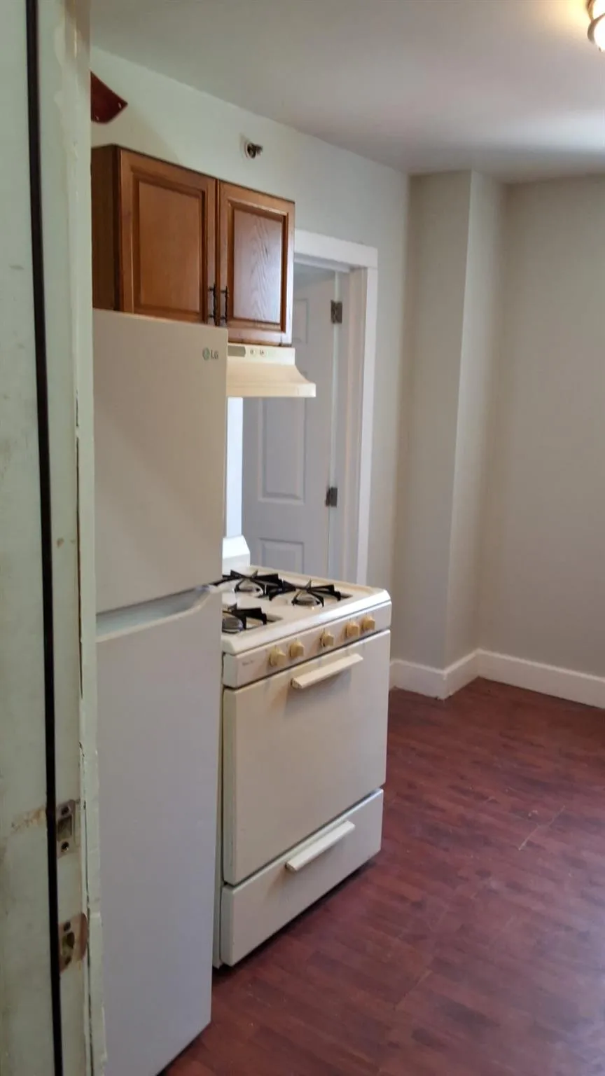 Kitchen featuring brown cabinets, white appliances, dark wood finished floors, and under cabinet range hood Kitchen featuring brown cabinets, white appliances, dark wood finished floors, and under cabinet range hood