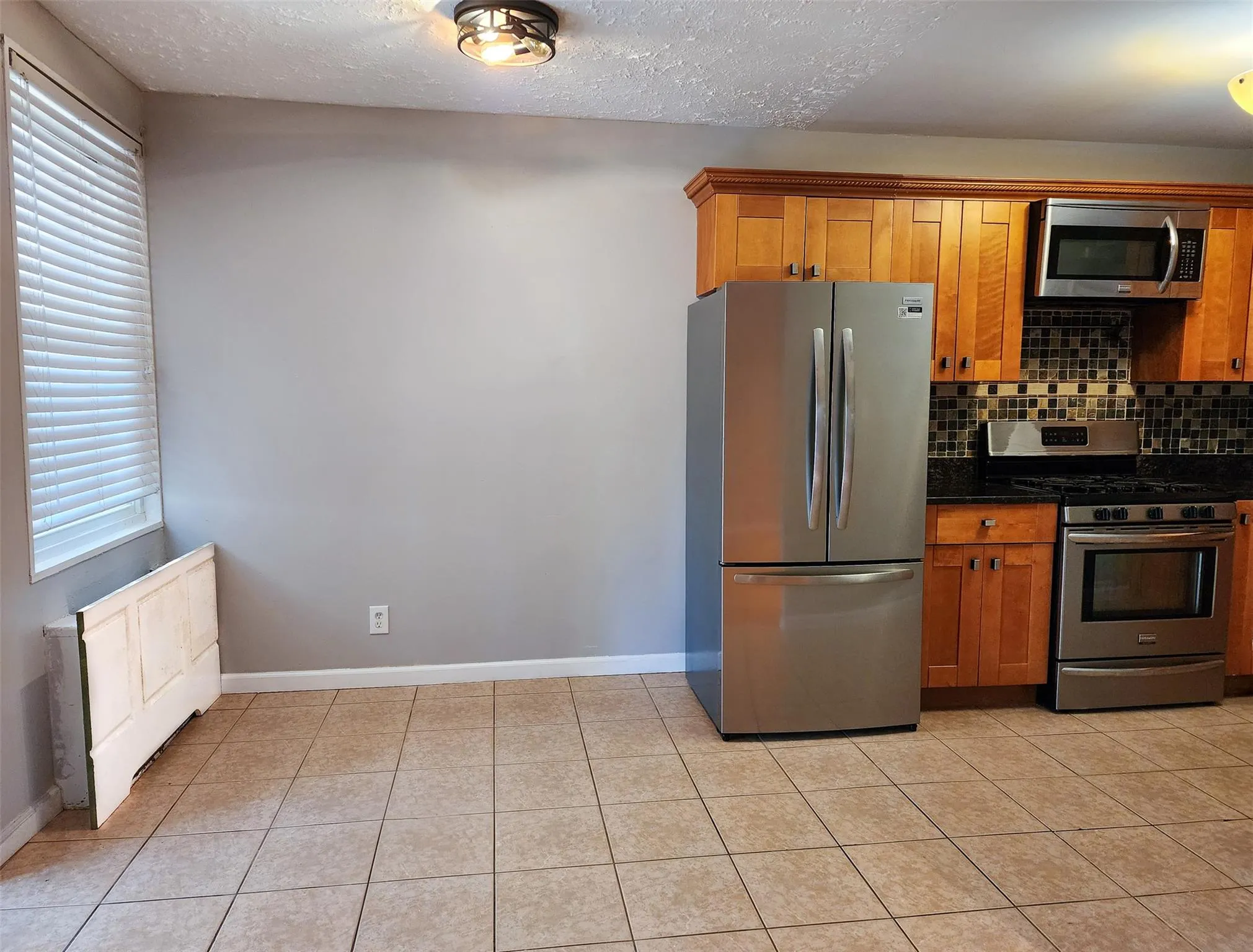 Kitchen featuring appliances with stainless steel finishes, decorative backsplash, dark countertops, brown cabinetry, and a textured ceiling Kitchen featuring appliances with stainless steel finishes, decorative backsplash, dark countertops, brown cabinetry, and a textured ceiling