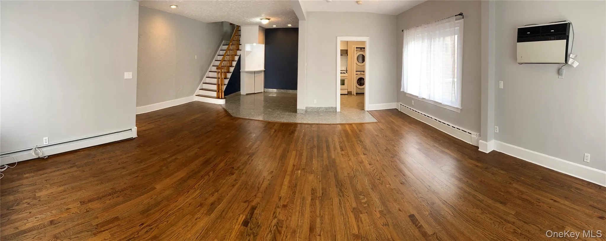 Unfurnished living room featuring stacked washer / dryer, a baseboard radiator, stairway, a textured ceiling, and wood finished floors Unfurnished living room featuring stacked washer / dryer, a baseboard radiator, stairway, a textured ceiling, and wood finished floors