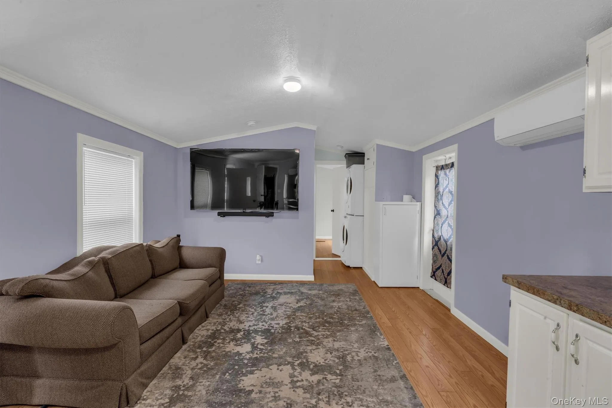 Living room featuring estacked washer and dryer, crown molding, light wood finished floors, vaulted ceiling, and a wall unit AC Living room featuring estacked washer and dryer, crown molding, light wood finished floors, vaulted ceiling, and a wall unit AC