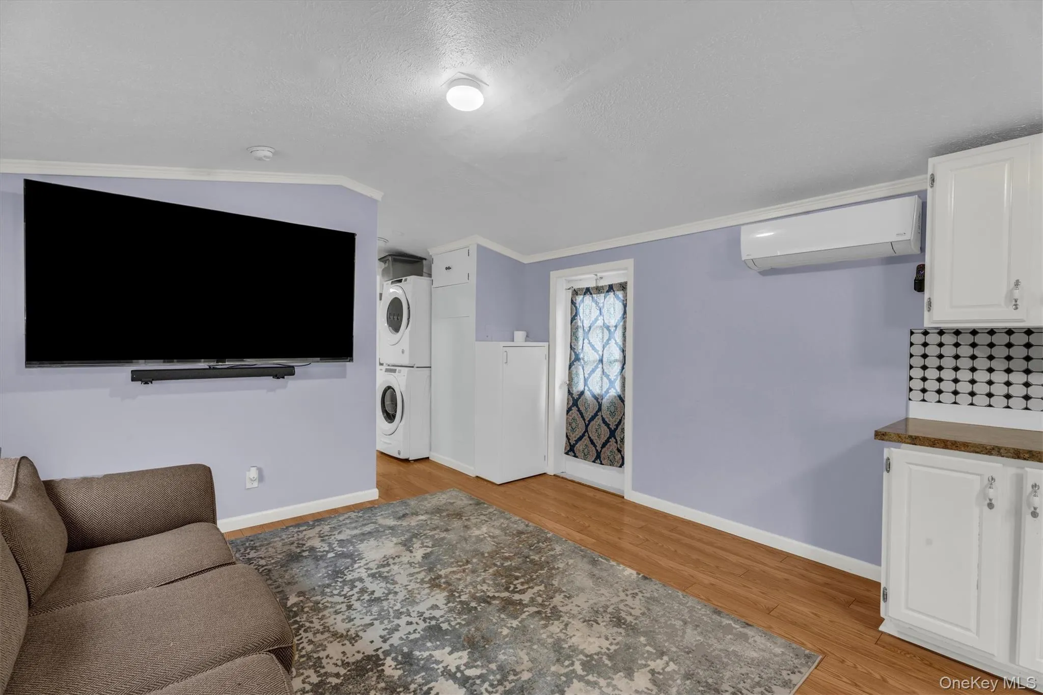Living area with estacked washer and dryer, ornamental molding, light wood finished floors, a wall mounted air conditioner, and a textured ceiling Living area with estacked washer and dryer, ornamental molding, light wood finished floors, a wall mounted air conditioner, and a textured ceiling