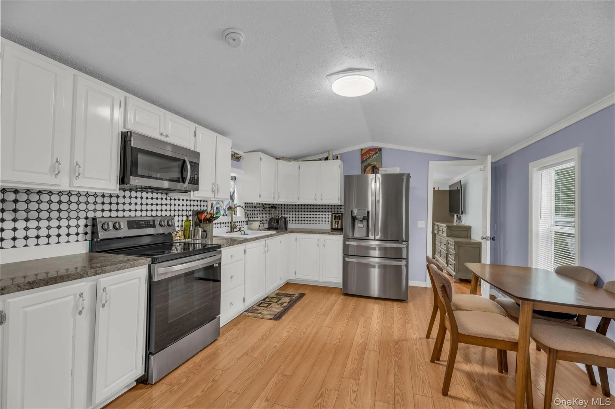 Kitchen with appliances with stainless steel finishes, backsplash, light wood-type flooring, vaulted ceiling, and white cabinetry Kitchen with appliances with stainless steel finishes, backsplash, light wood-type flooring, vaulted ceiling, and white cabinetry