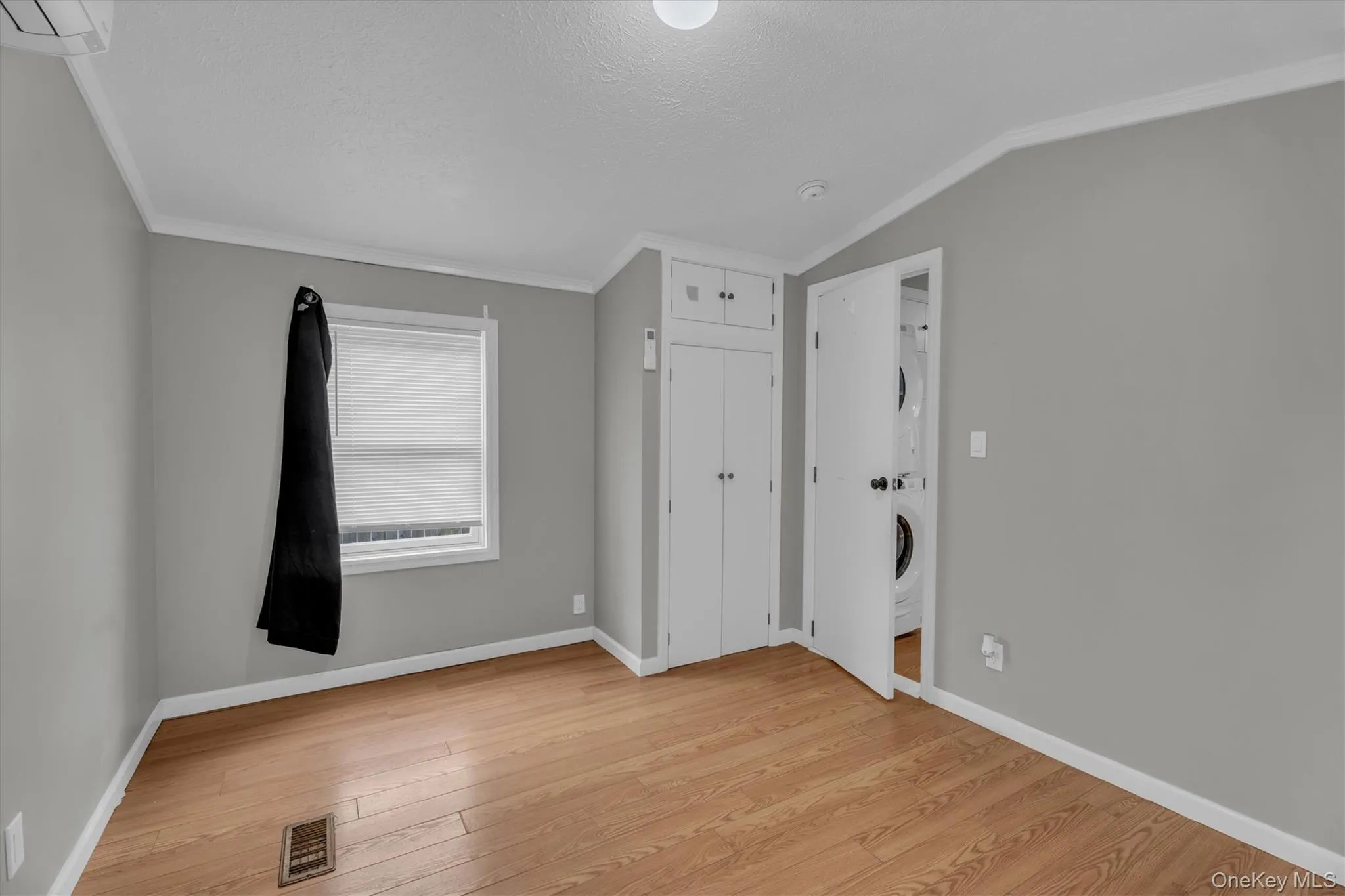 Unfurnished bedroom with ornamental molding, light wood finished floors, stacked washer and clothes dryer, a textured ceiling, and lofted ceiling Unfurnished bedroom with ornamental molding, light wood finished floors, stacked washer and clothes dryer, a textured ceiling, and lofted ceiling