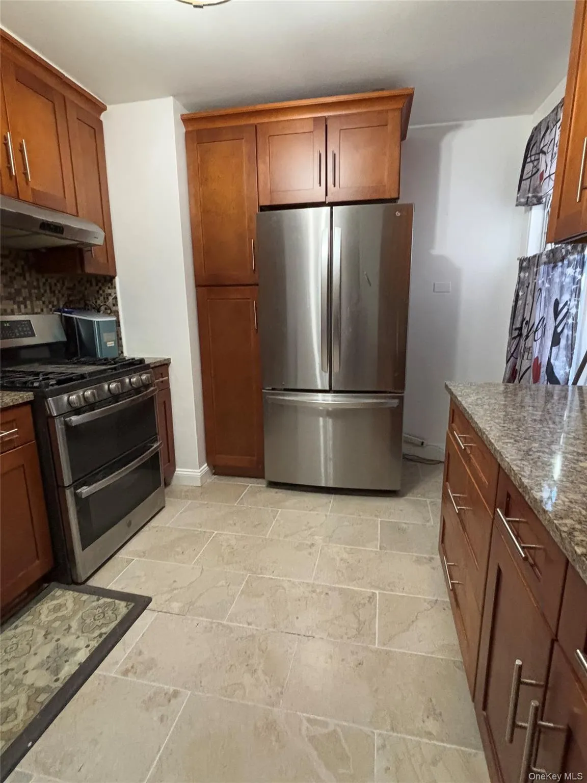 Kitchen with light stone counters, brown cabinets, stainless steel appliances, under cabinet range hood, and backsplash Kitchen with light stone counters, brown cabinets, stainless steel appliances, under cabinet range hood, and backsplash