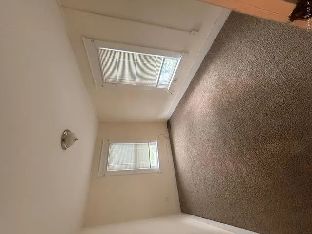 Carpeted interior space featuring two windows with blinds, a ceiling-mounted light fixture, and neutral-toned walls Carpeted interior space featuring two windows with blinds, a ceiling-mounted light fixture, and neutral-toned walls