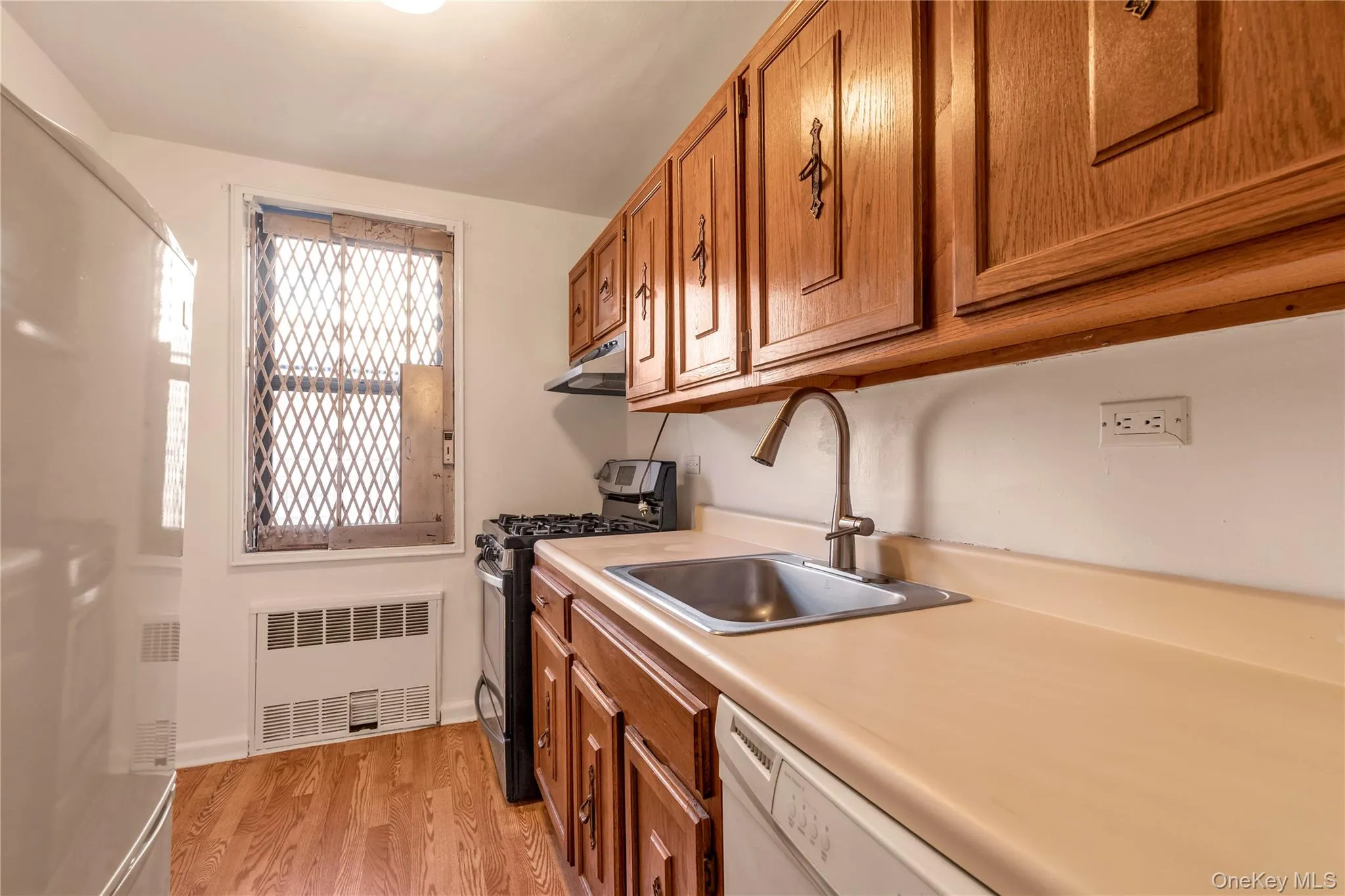Kitchen with brown cabinetry, white appliances, radiator heating unit, light countertops, and light wood-type flooring Kitchen with brown cabinetry, white appliances, radiator heating unit, light countertops, and light wood-type flooring