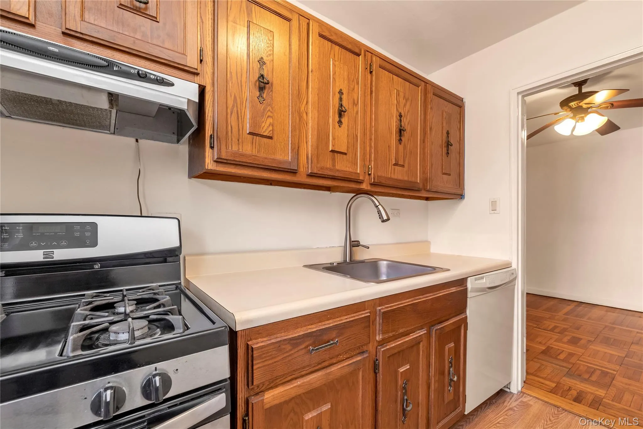 Kitchen featuring stainless steel gas range oven, exhaust hood, brown cabinetry, white dishwasher, and light countertops Kitchen featuring stainless steel gas range oven, exhaust hood, brown cabinetry, white dishwasher, and light countertops