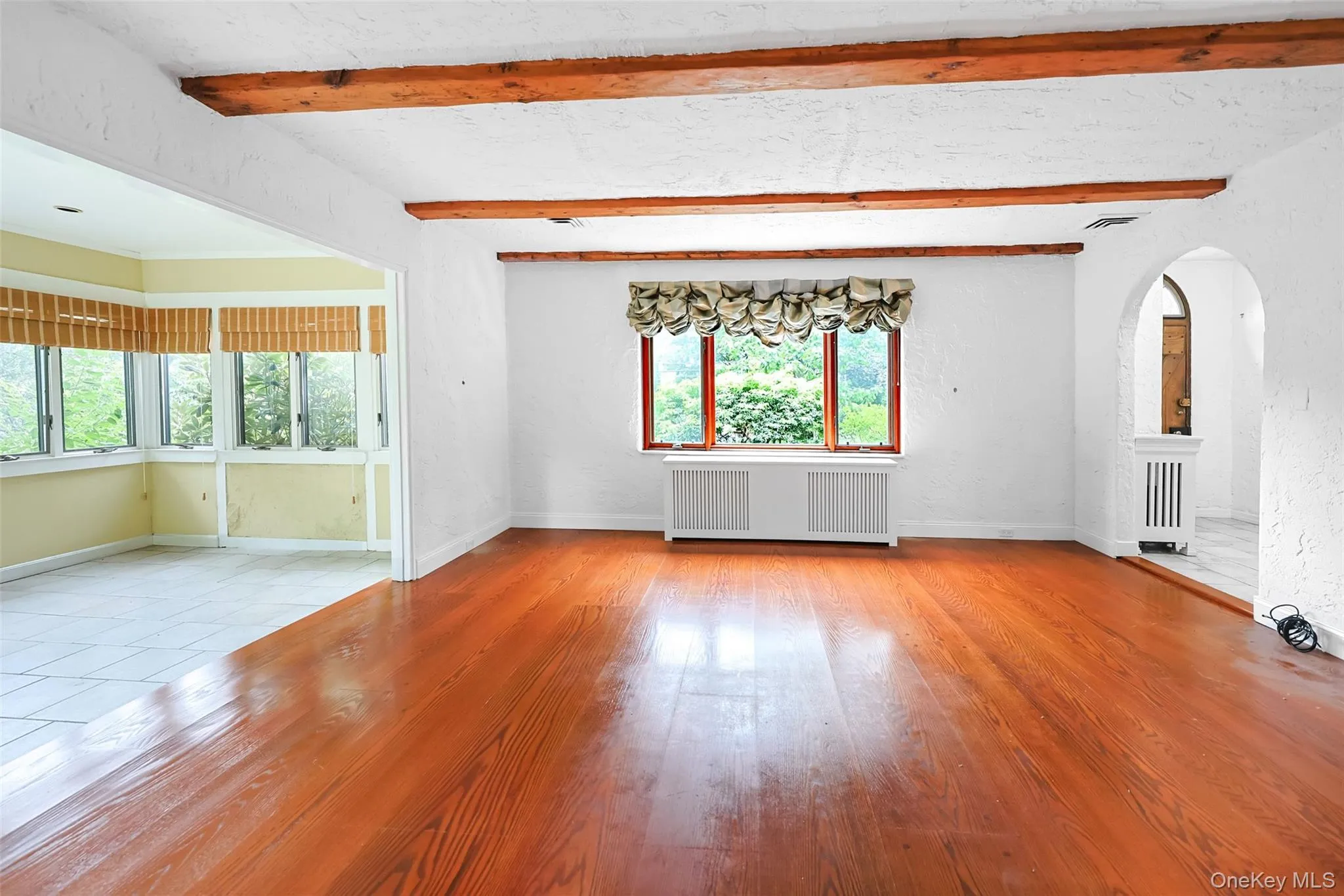 Unfurnished living room with beamed ceiling, radiator, arched walkways, and wood finished floors Unfurnished living room with beamed ceiling, radiator, arched walkways, and wood finished floors