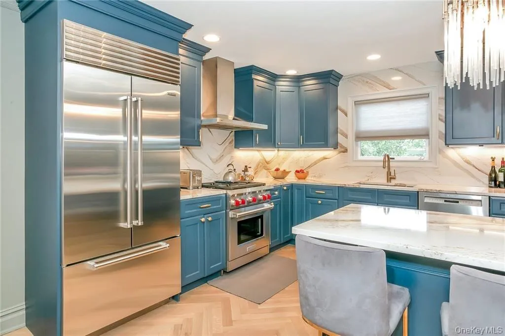 Kitchen with blue cabinetry, premium appliances, wall chimney exhaust hood, tasteful backsplash, and light stone countertops Kitchen with blue cabinetry, premium appliances, wall chimney exhaust hood, tasteful backsplash, and light stone countertops