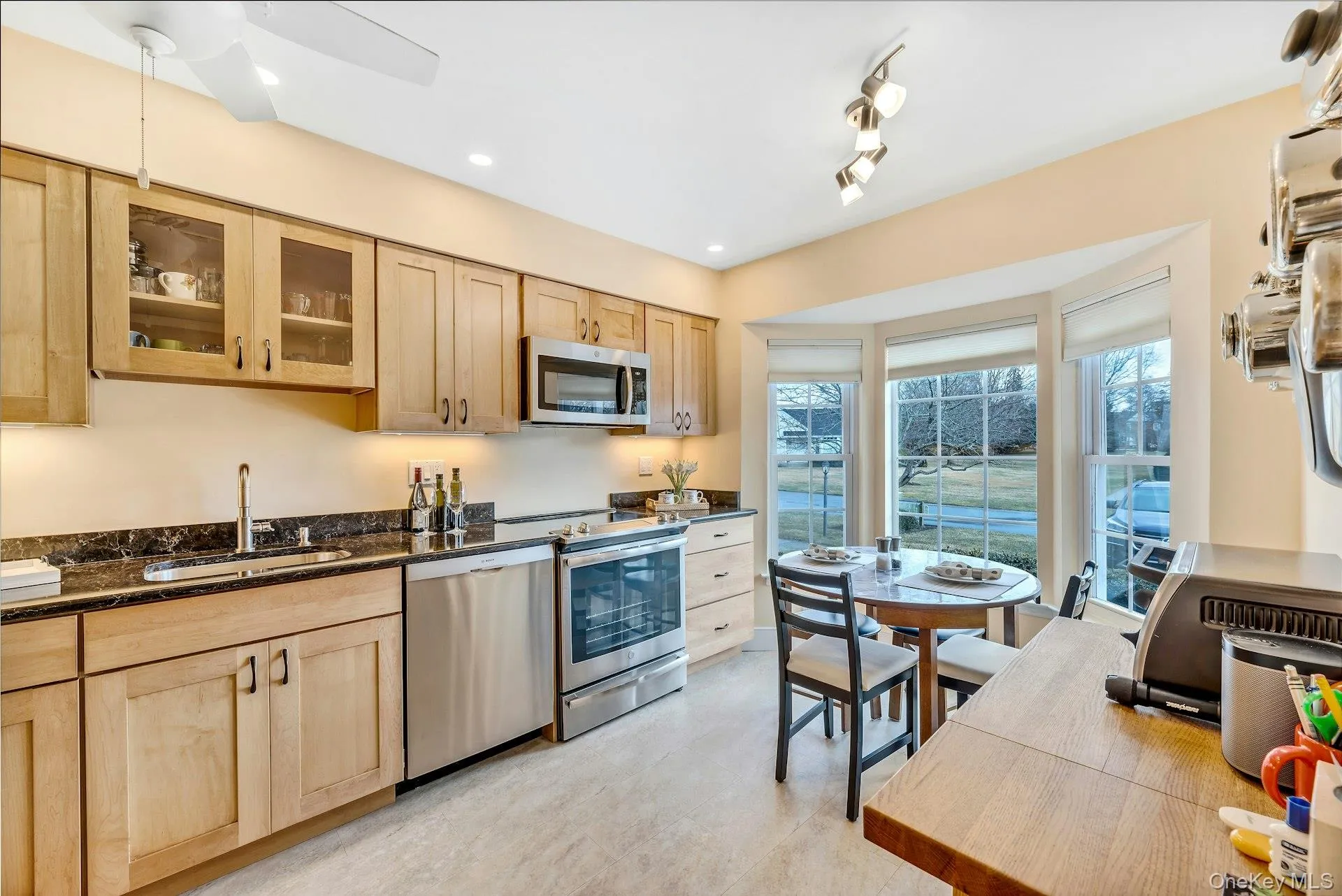 Kitchen featuring light brown cabinetry, dark stone counters, appliances with stainless steel finishes, glass insert cabinets, and recessed lighting Kitchen featuring light brown cabinetry, dark stone counters, appliances with stainless steel finishes, glass insert cabinets, and recessed lighting